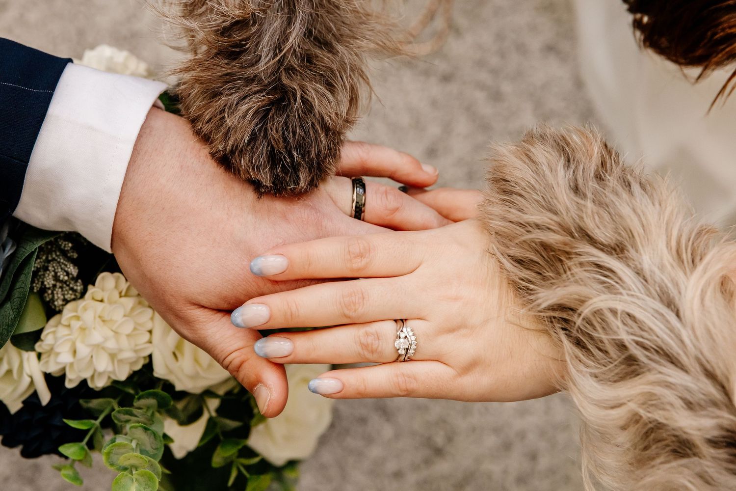 Newlywed couple holding hands showing wedding rings with bouquet and fluffy dog. The Grand Ivory