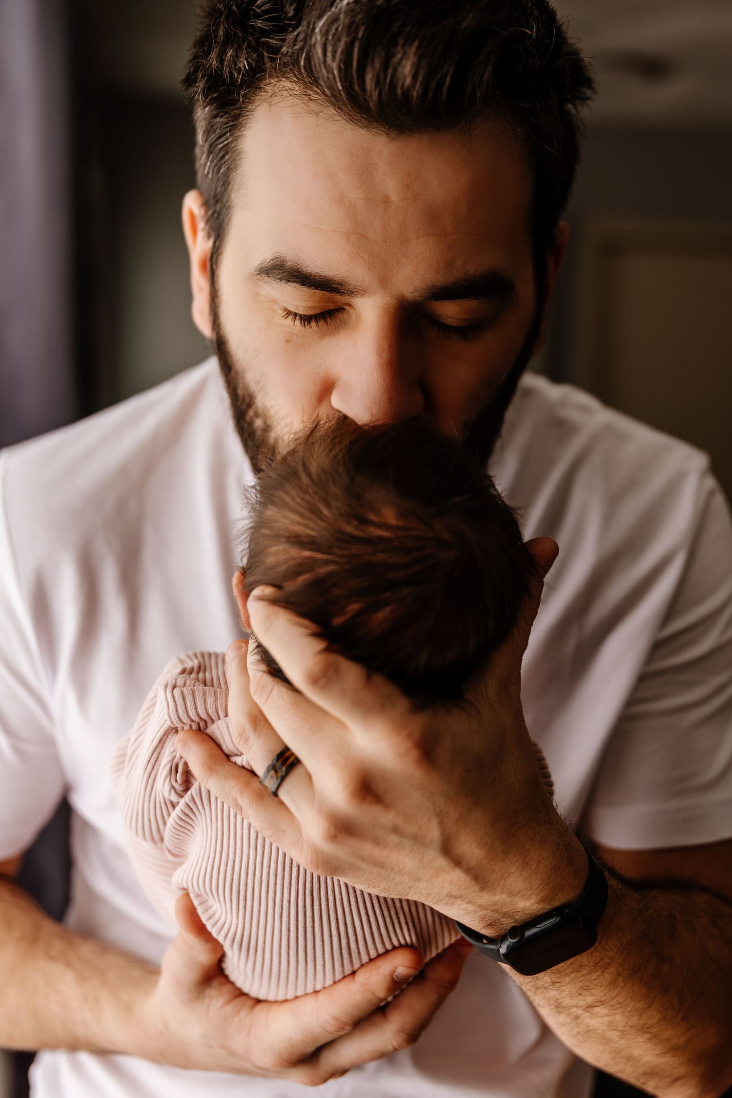 Bearded father in white t-shirt tenderly kissing his newborn baby's head.