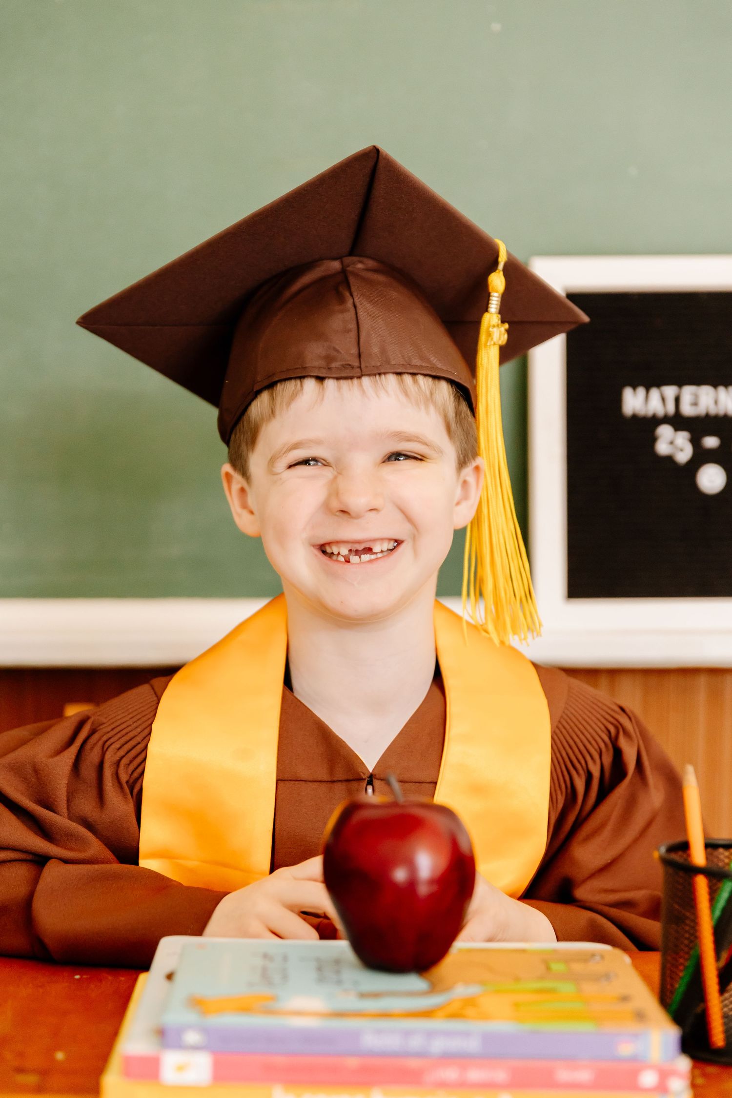 A smiling boy in a brown graduation cap and gown sits at a desk with an apple and books.