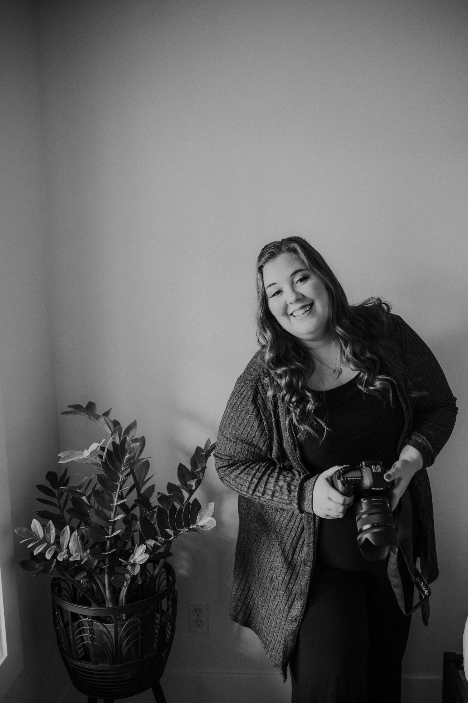 Thalie photography Black and white portrait of smiling female photographer holding DSLR camera next to indoor plant.
