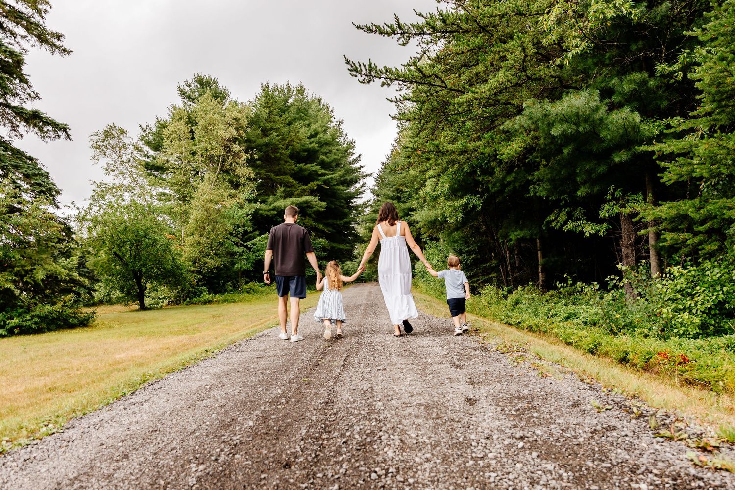 Family of four walking away down a tree-lined gravel road on a cloudy summer day.