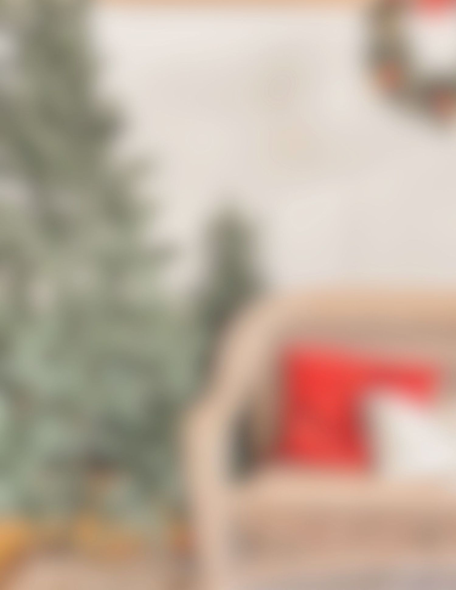A cozy Christmas living room with an undecorated green tree, wicker chair with red pillow, and wreath on white wall.
