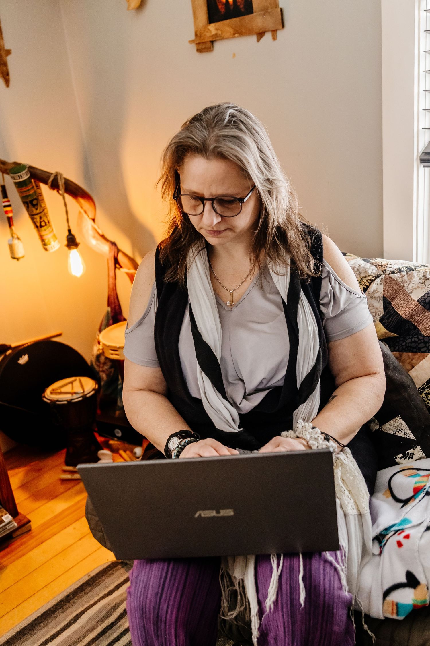 Woman with gray hair and glasses working on an ASUS laptop while sitting on a couch in a cozy home.