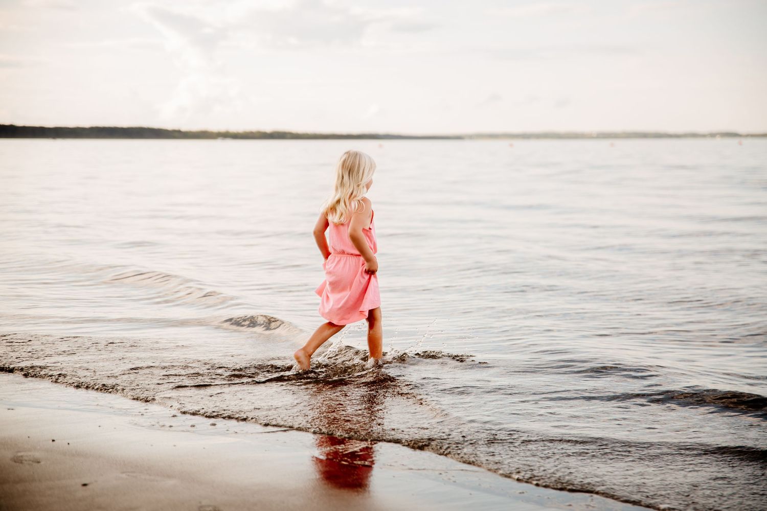 A young blonde girl in a pink dress walks along the shoreline, splashing in gentle ocean waves at sunset.