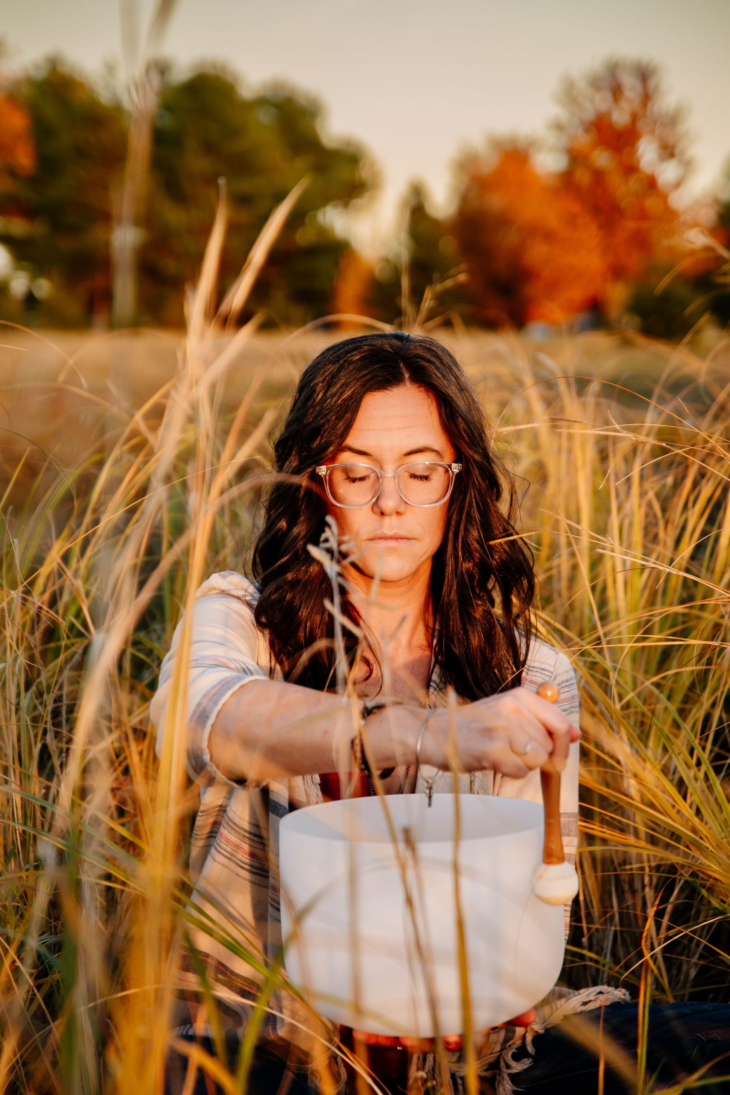 Woman with glasses playing a crystal singing bowl in a golden autumn grass field at sunset.