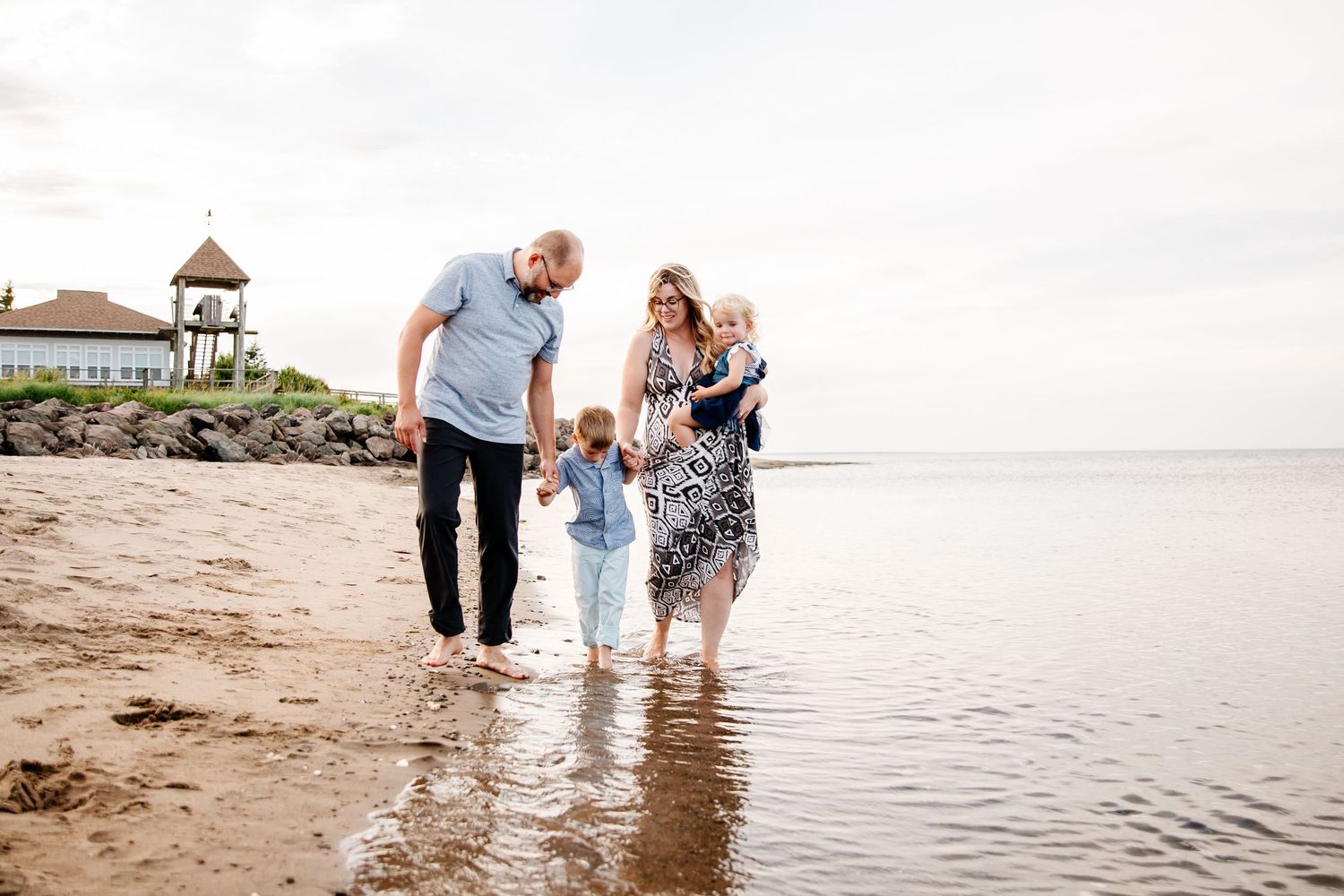 Family of four walking barefoot along a sandy beach shore, with a gazebo and rocky coastline in the background.