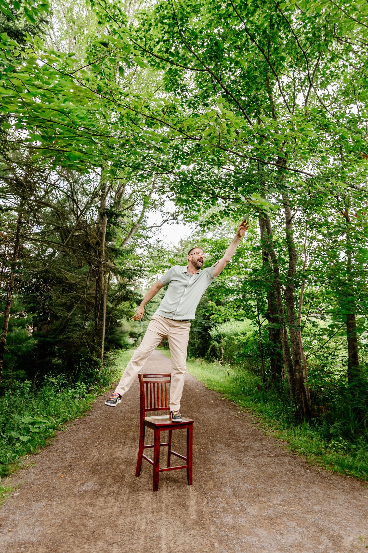 Man balancing on wooden chair on forest path with arms outstretched, surrounded by lush green trees.