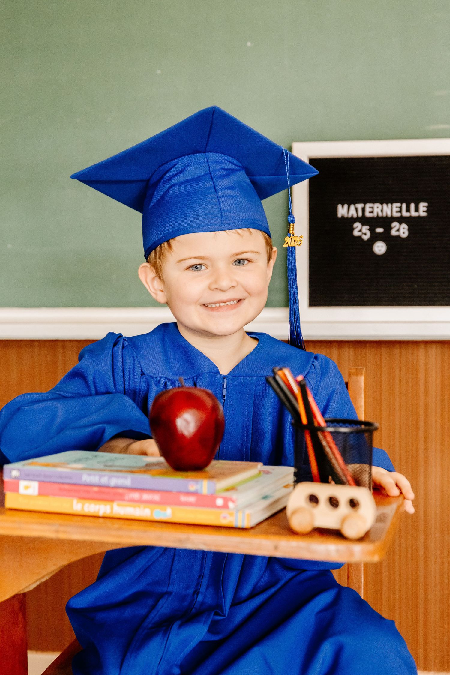 A cheerful young boy in a blue graduation cap and gown for kindergarten graduation sits at a school desk with books and an apple.