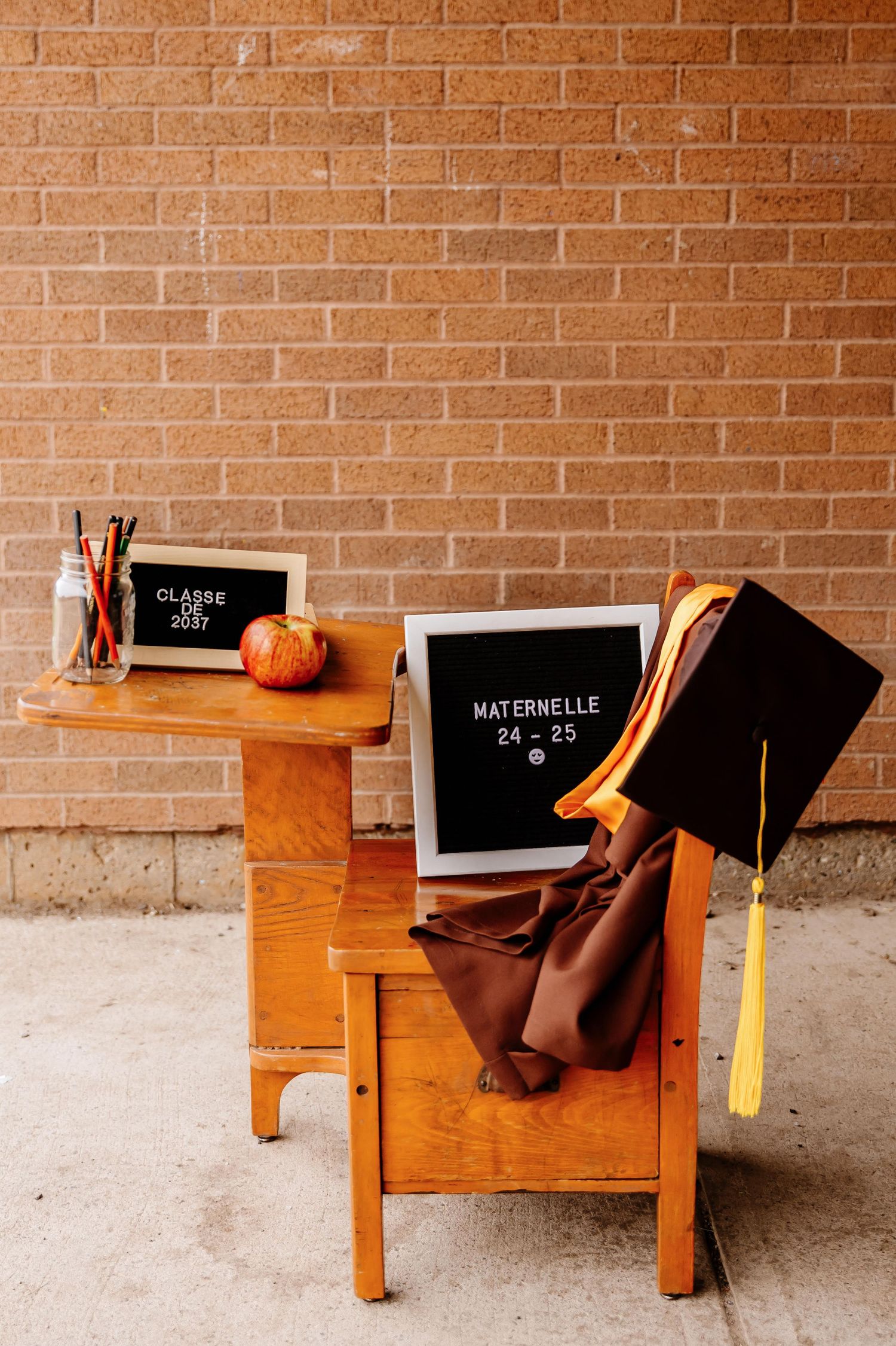Brown graduation cap and gown with Maternelle 24-25 sign displayed on a wooden school desk outdoors.