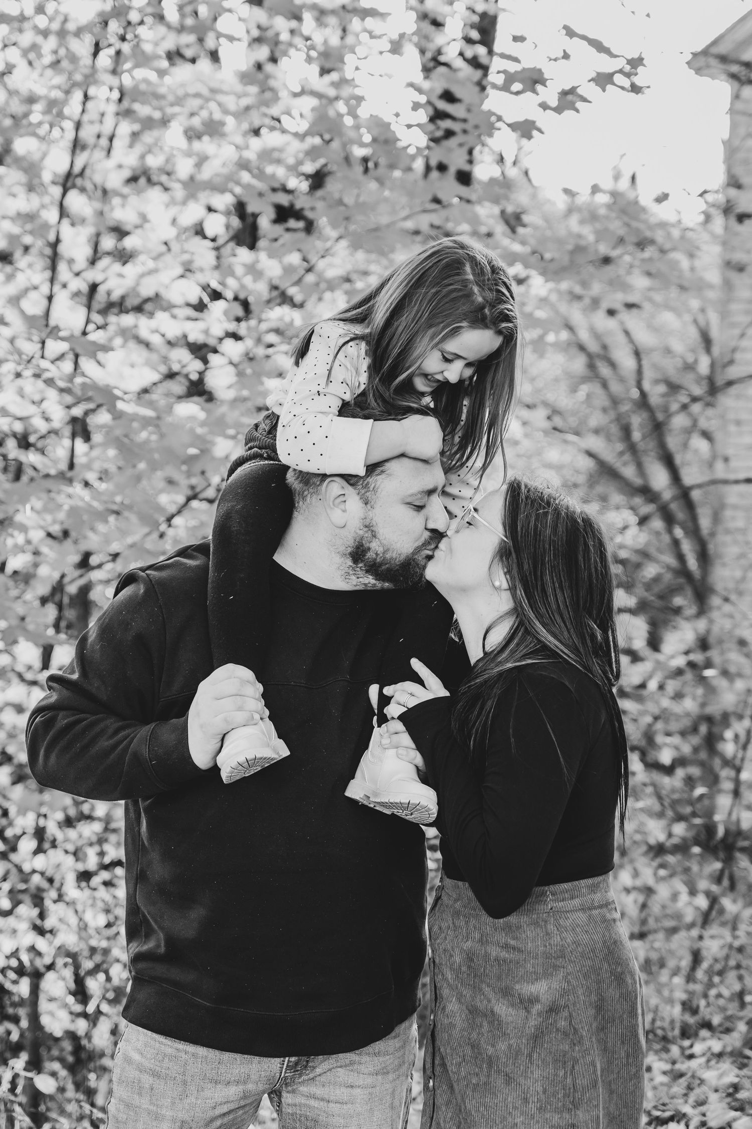 Black and white photo of couple kissing while daughter sits on fathers shoulders outdoors. Rogersville