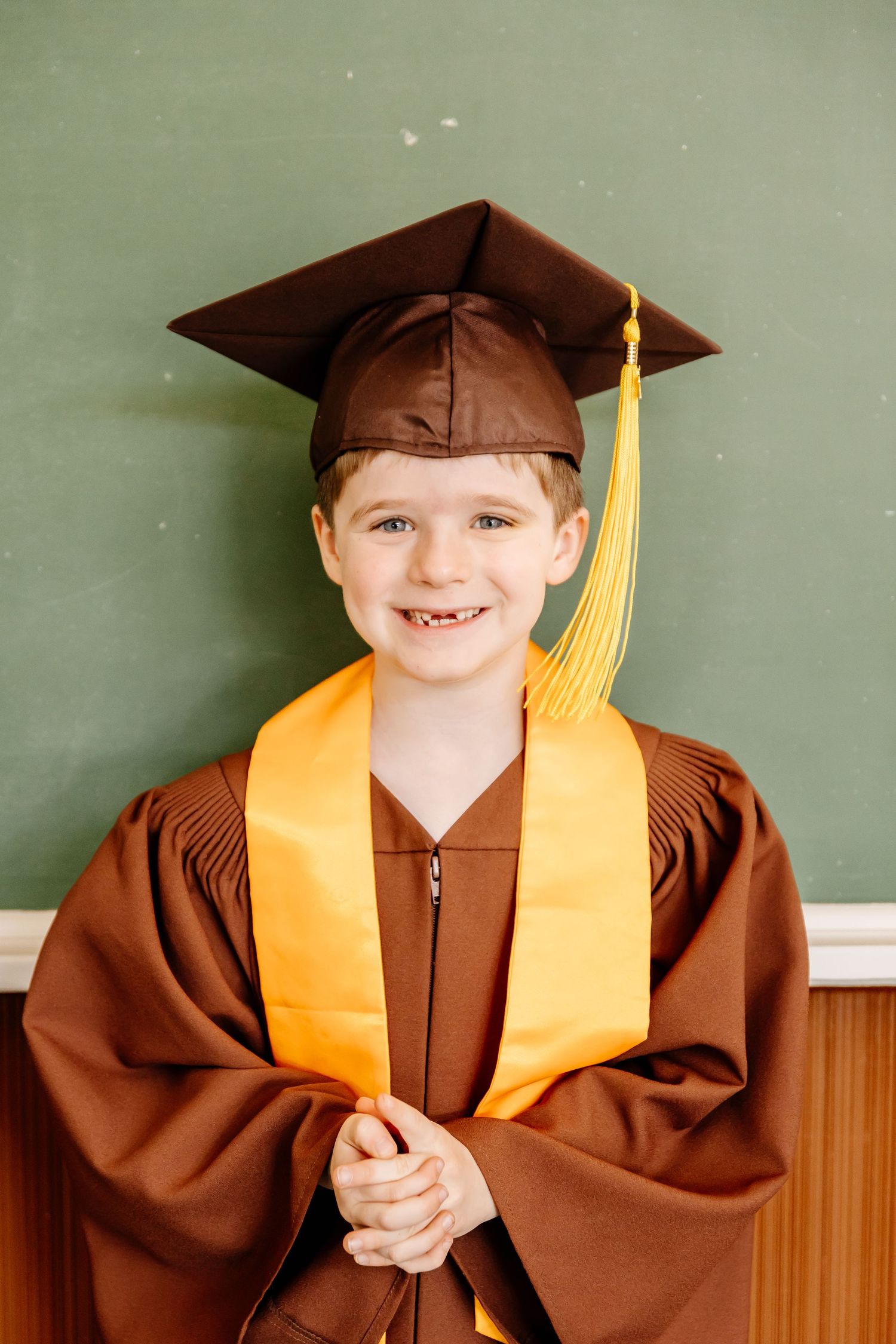 A smiling young boy in a brown graduation cap and gown for LJR school stands in front of a green chalkboard for it's kingergarten graduation
