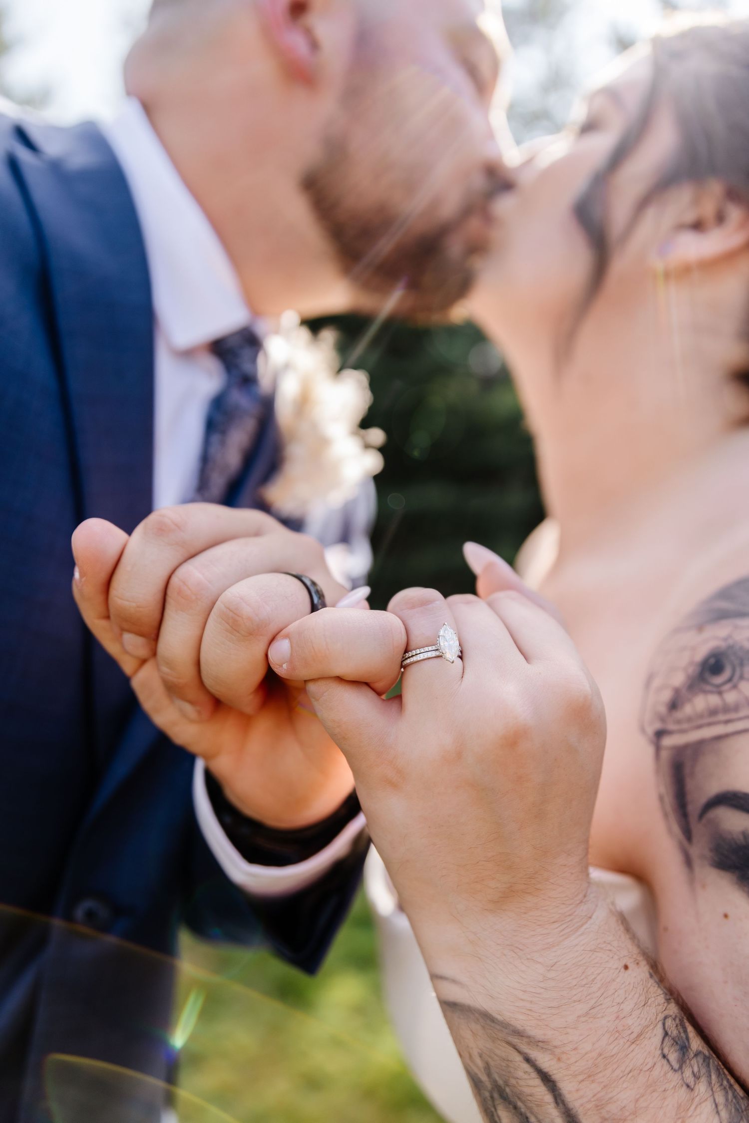 Newlyweds kissing while holding hands showing wedding rings, bride with tattoos wearing diamond engagement ring.