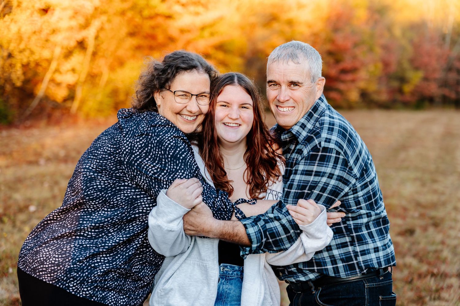 Family of three smiling outdoors in autumn, embracing warmly with colorful fall foliage in background.