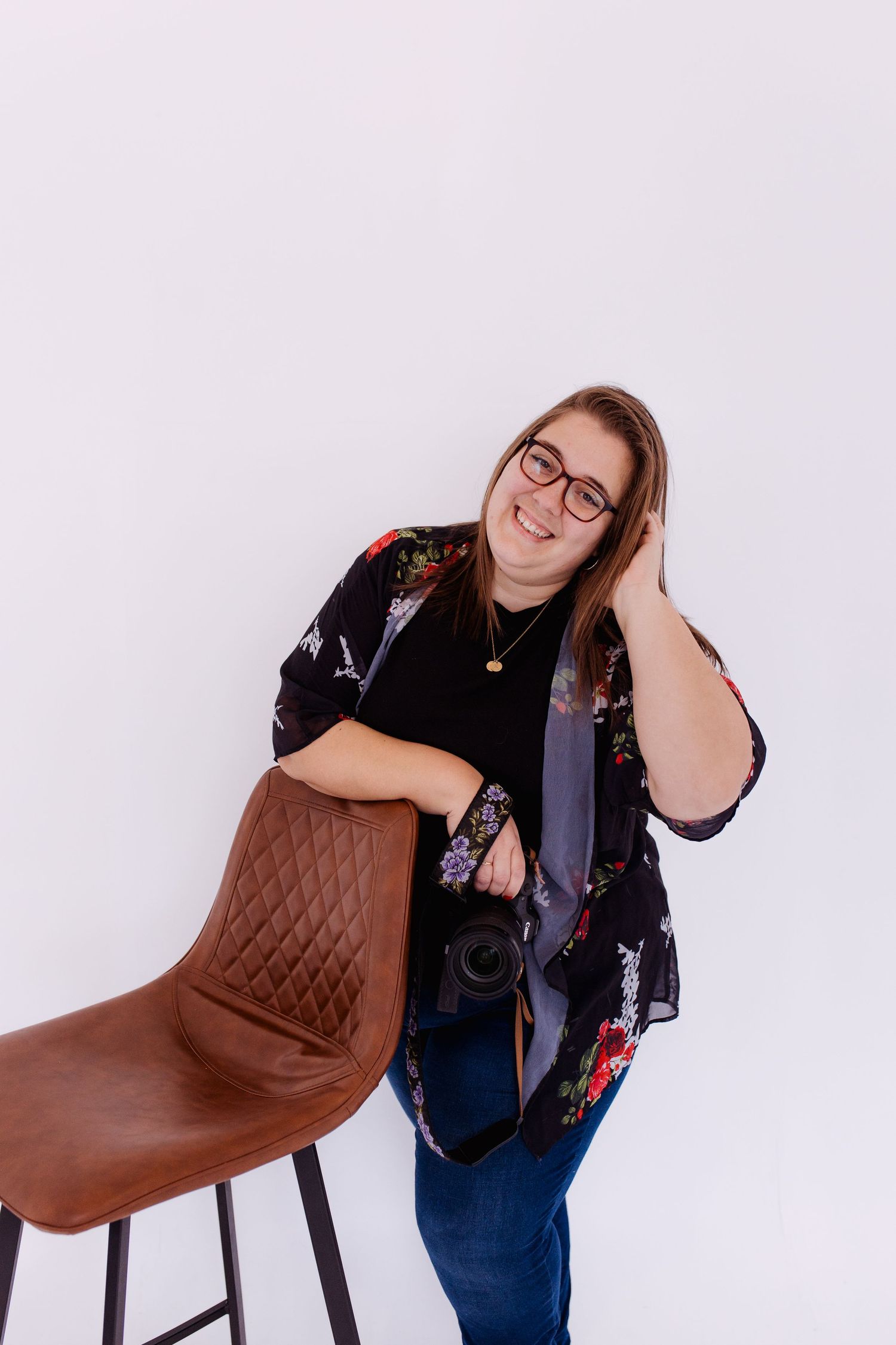 Thalie photography Smiling female photographer in floral kimono leaning on brown leather chair holding camera in white studio.
