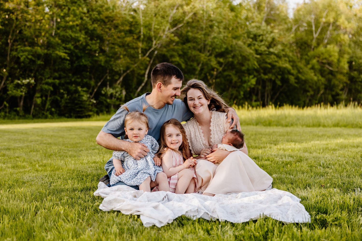 Happy family of five sits together on white blanket in green meadow during golden hour.