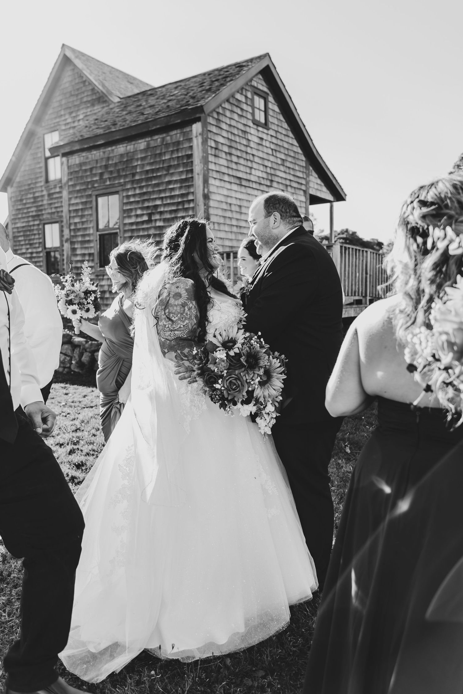 Black and white photo of bride and groom walking with guests outside a rustic barn.