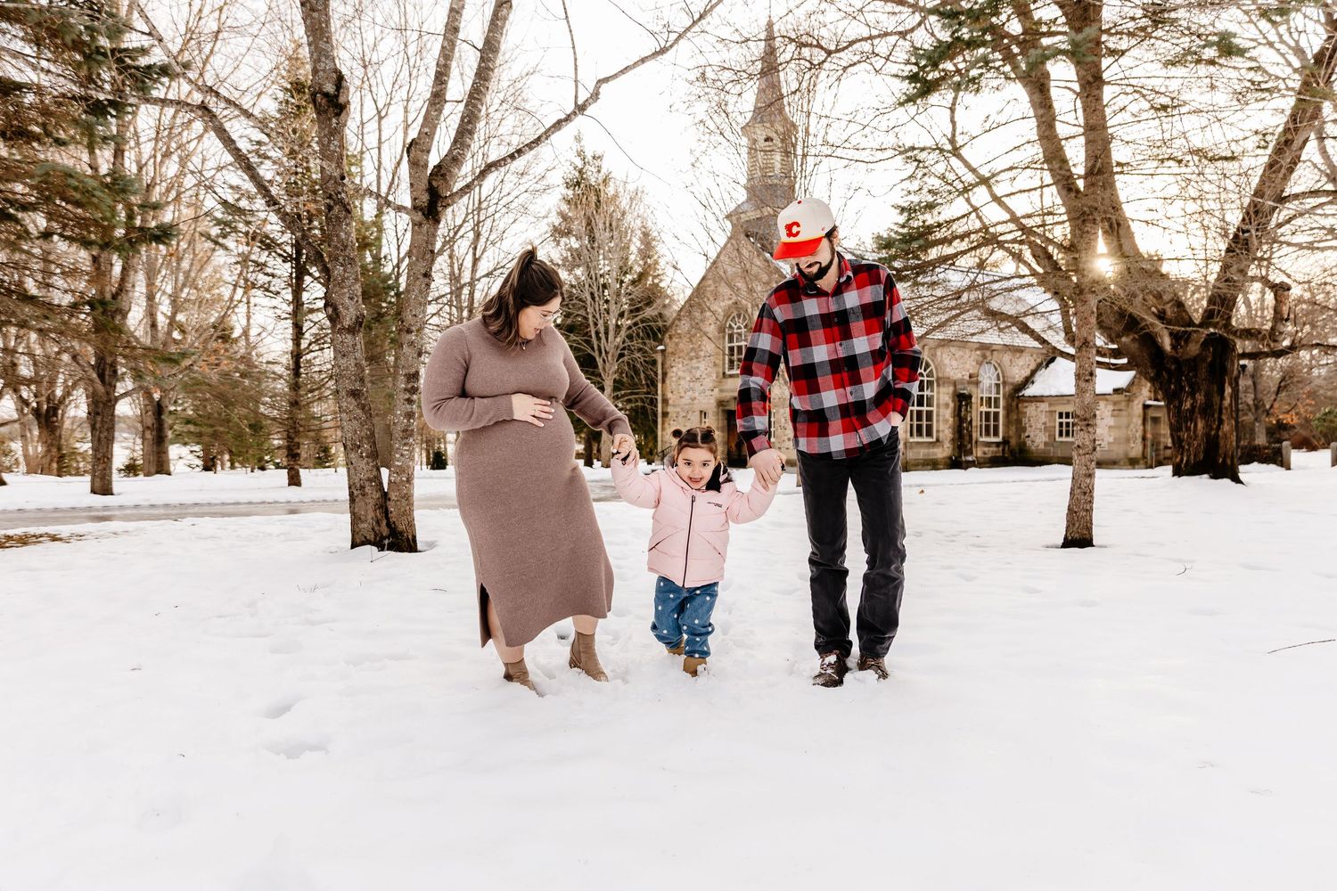 Pregnant couple walking hand-in-hand with their toddler through a snowy winter landscape.