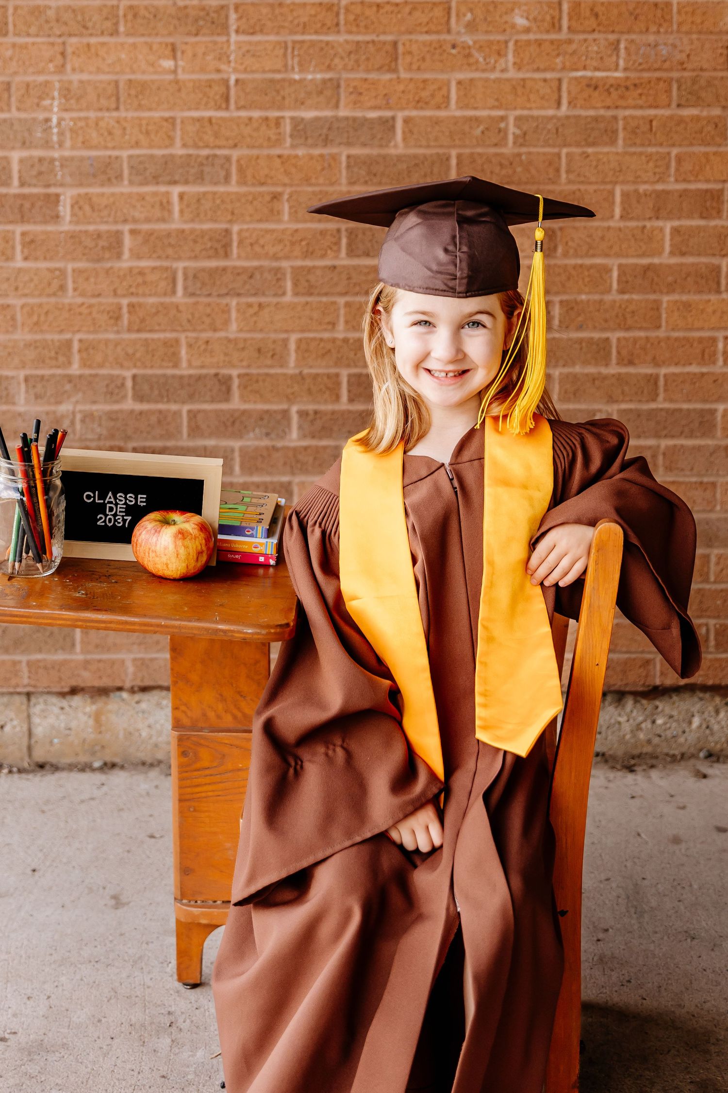 Smiling young girl in brown and gold graduation cap and gown seated at a wooden school desk.