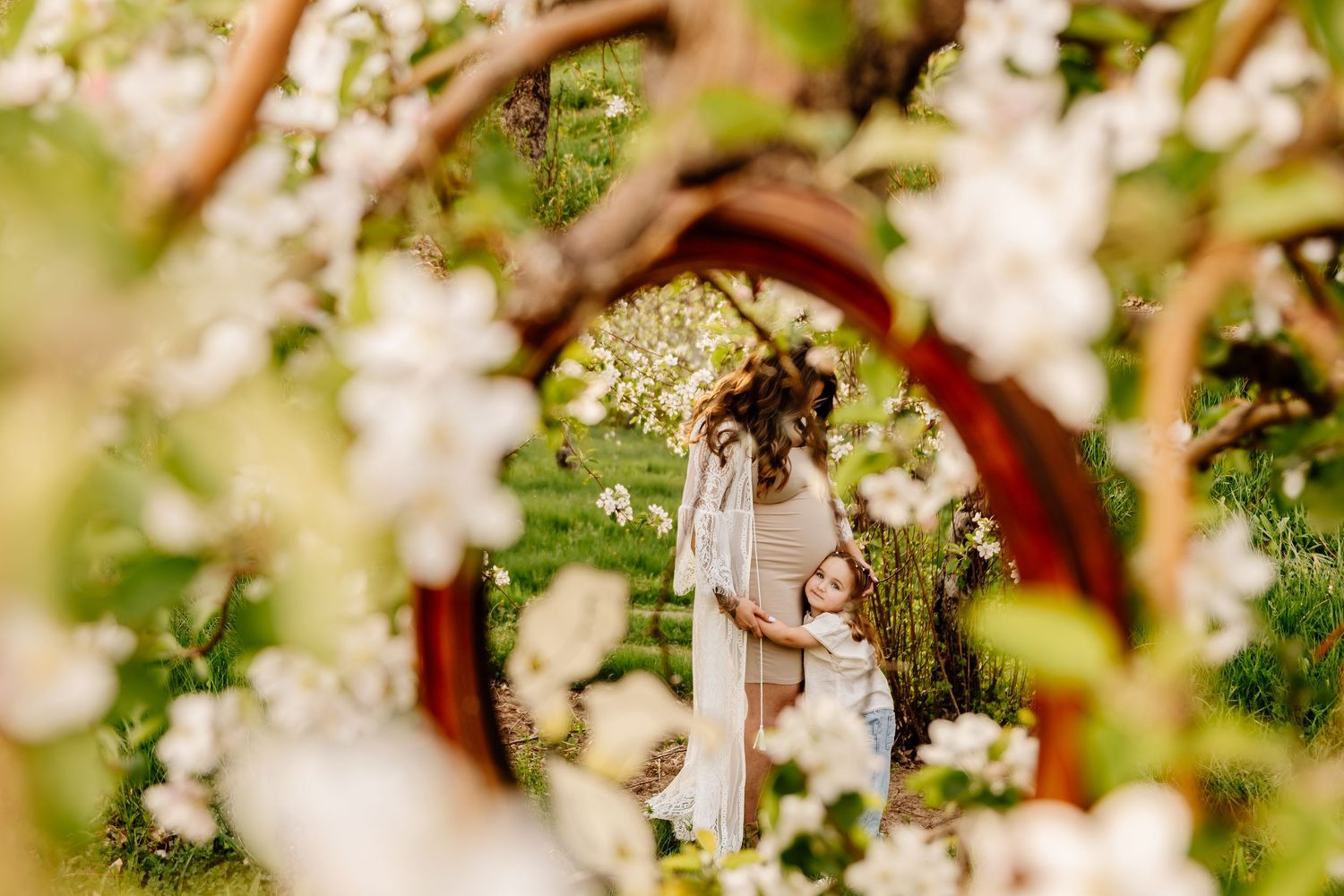 Mother and daughter in white dresses standing under a floral arch surrounded by blooming white spring flowers.