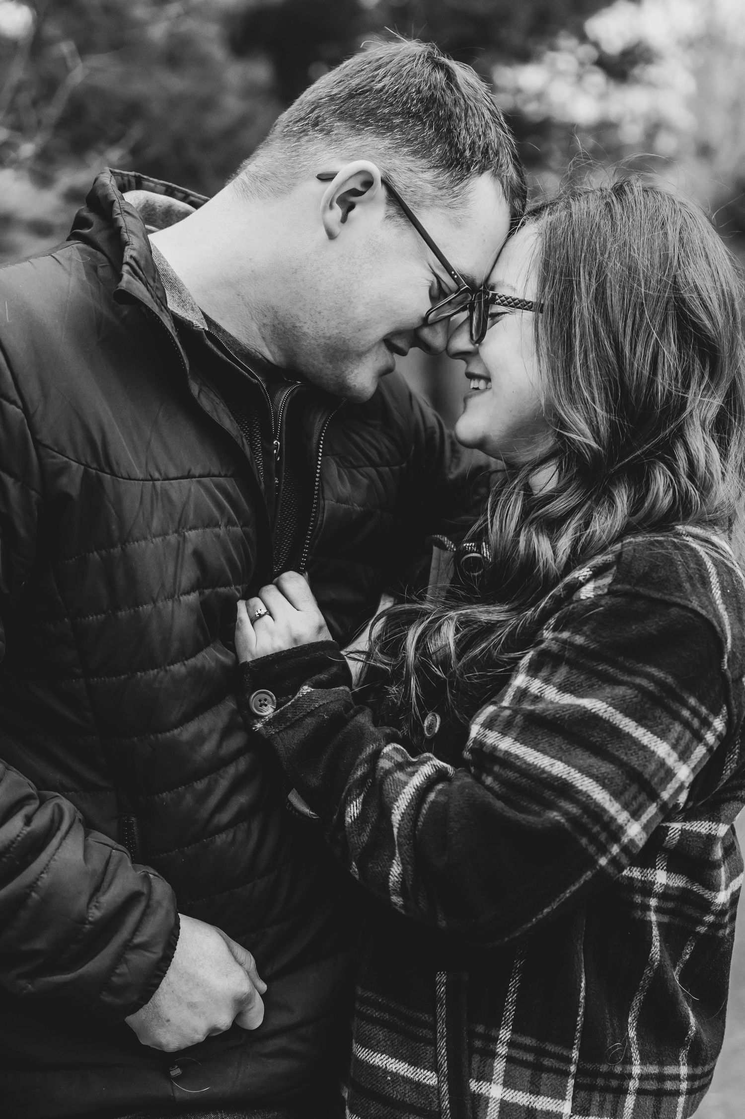 Black and white portrait of a smiling couple in glasses sharing a tender forehead touch outdoors.