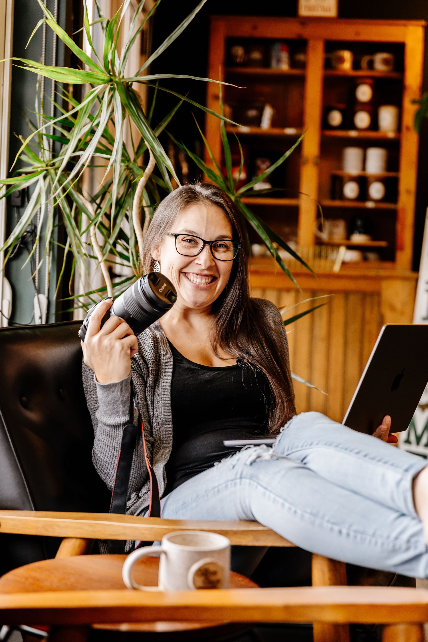 A smiling woman with glasses sits in a chair holding a camera with a laptop and coffee cup nearby.