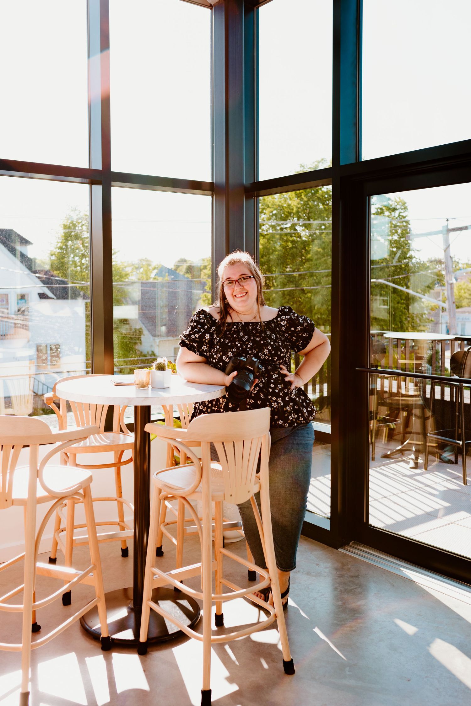 Thalie photography Smiling blonde woman in floral top leaning on high-top table in sunlit modern cafe with large windows.