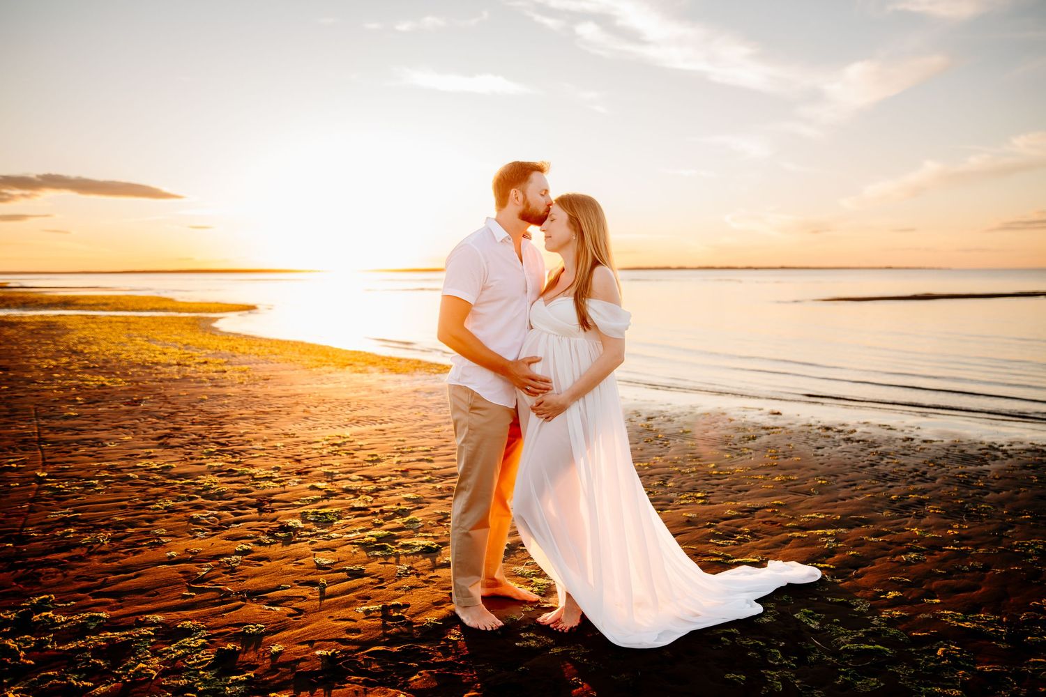 Expecting couple embracing at sunset on a beach, with the woman wearing a flowing white gown.