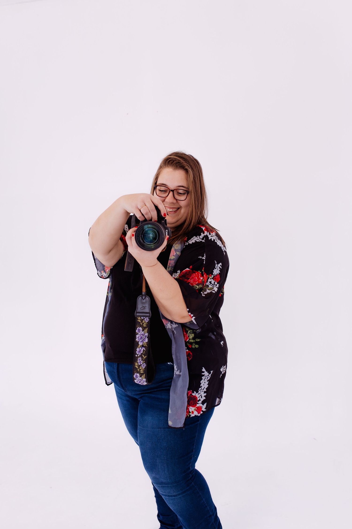 Thalie photography Female photographer in floral kimono pointing DSLR camera toward viewer in bright white photography studio.