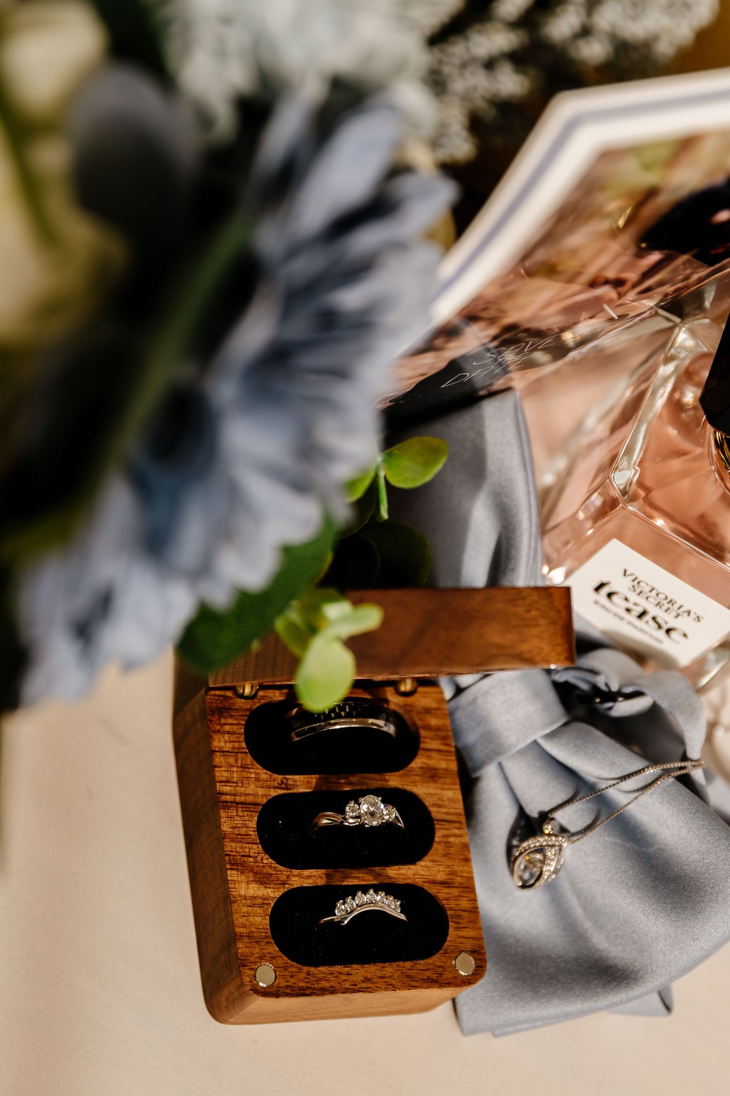 Elegant wedding rings displayed in a wooden velvet box alongside perfume and blue flowers.