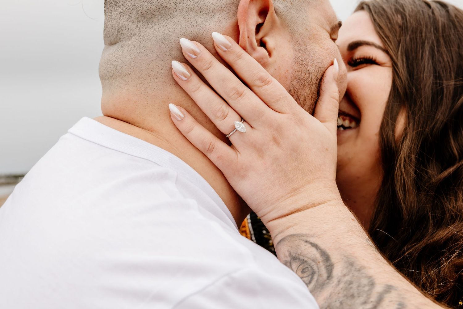 Close-up of laughing couple embracing, showcasing engagement ring on brides hand with tattooed groom.