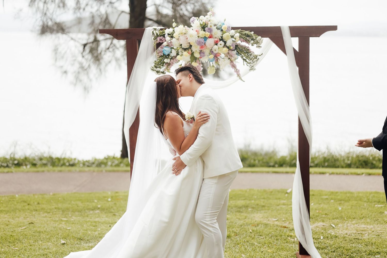 Bride and groom share first kiss under floral arch at outdoor waterfront wedding ceremony.