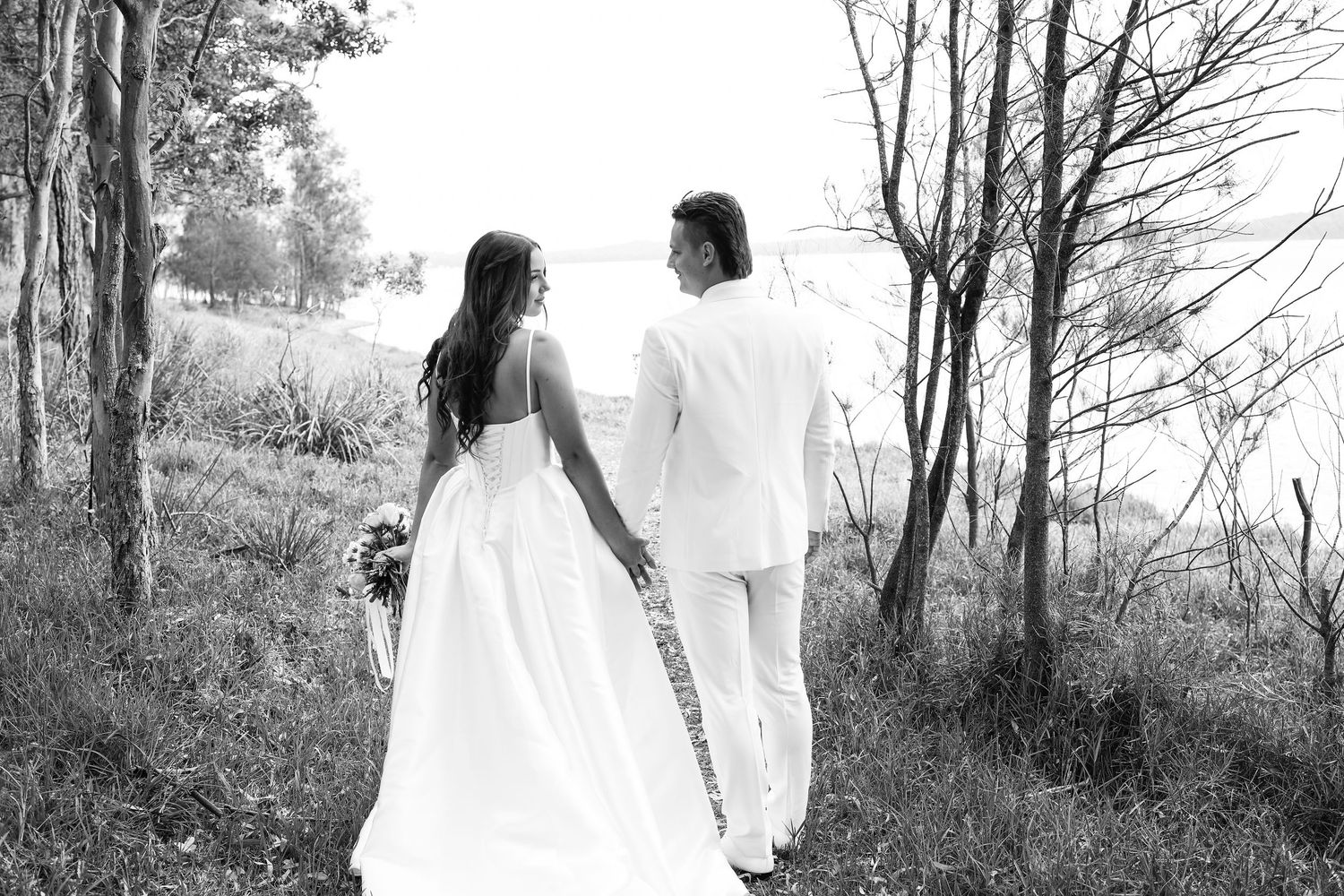 Black and white photo of bride and groom walking hand in hand through forest path.