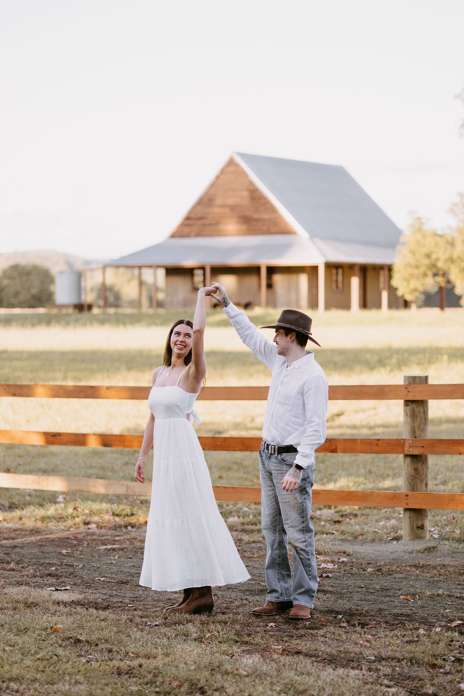 Couple dancing joyfully near a wooden fence with a rustic barn and open fields in background.