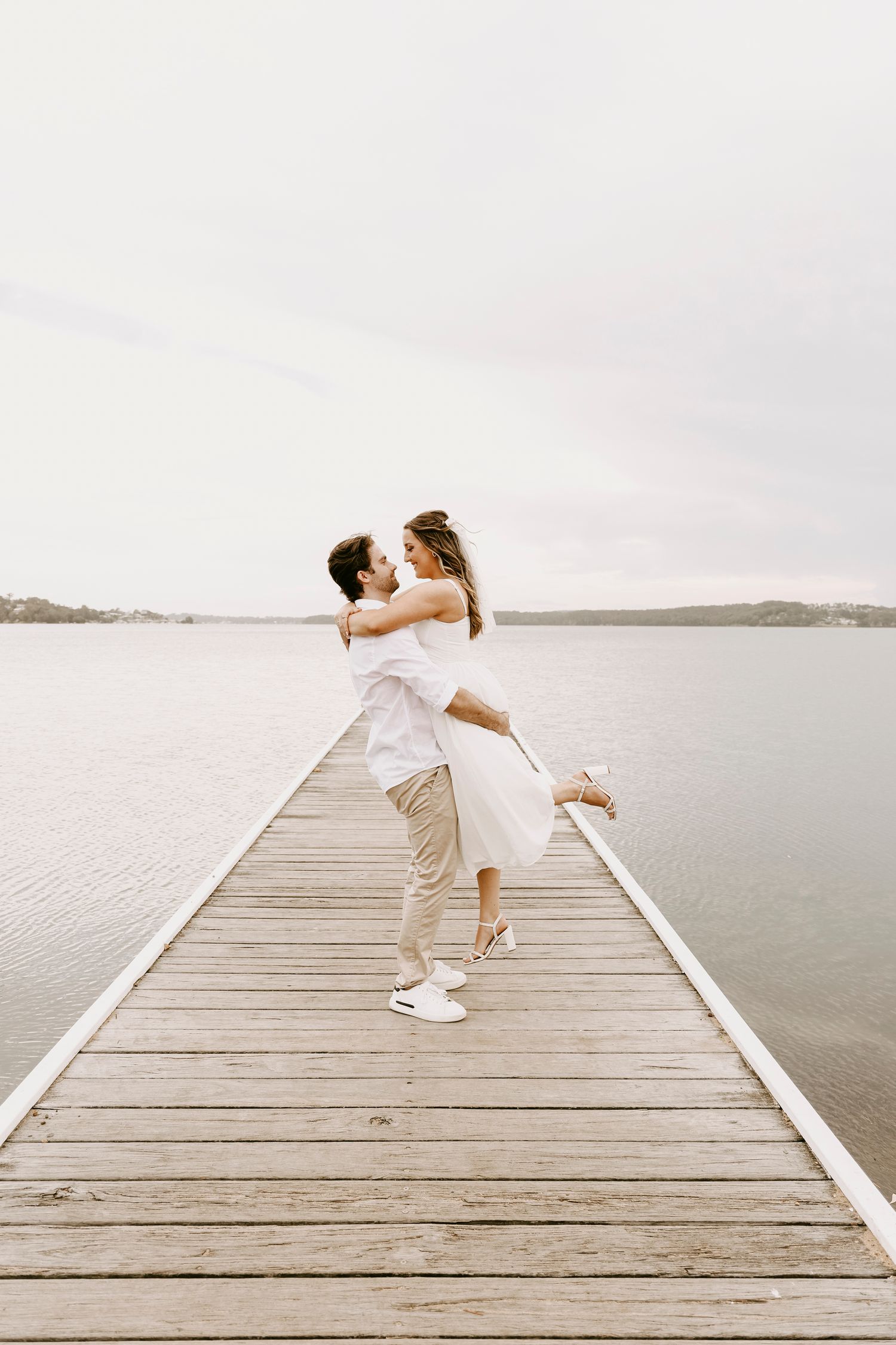Groom lifting bride on a wooden pier with serene lake backdrop on wedding day.