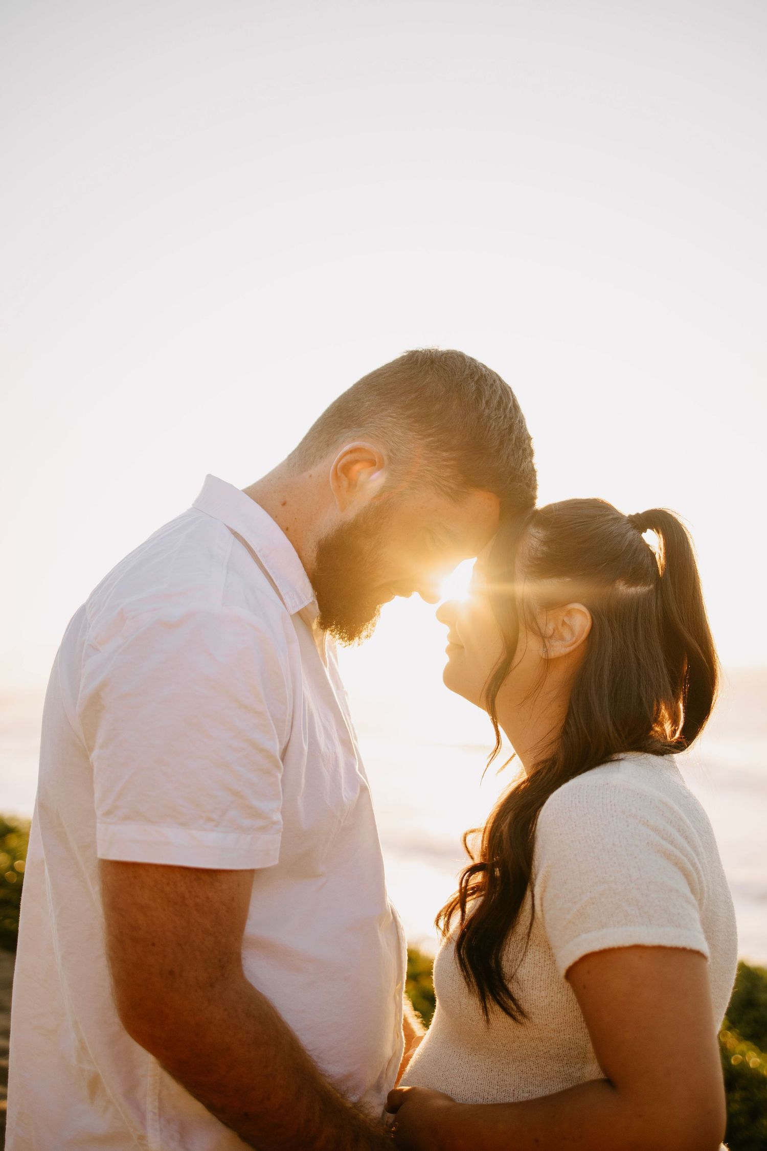 A couple touches foreheads romantically with golden sunset light glowing between them outdoors.