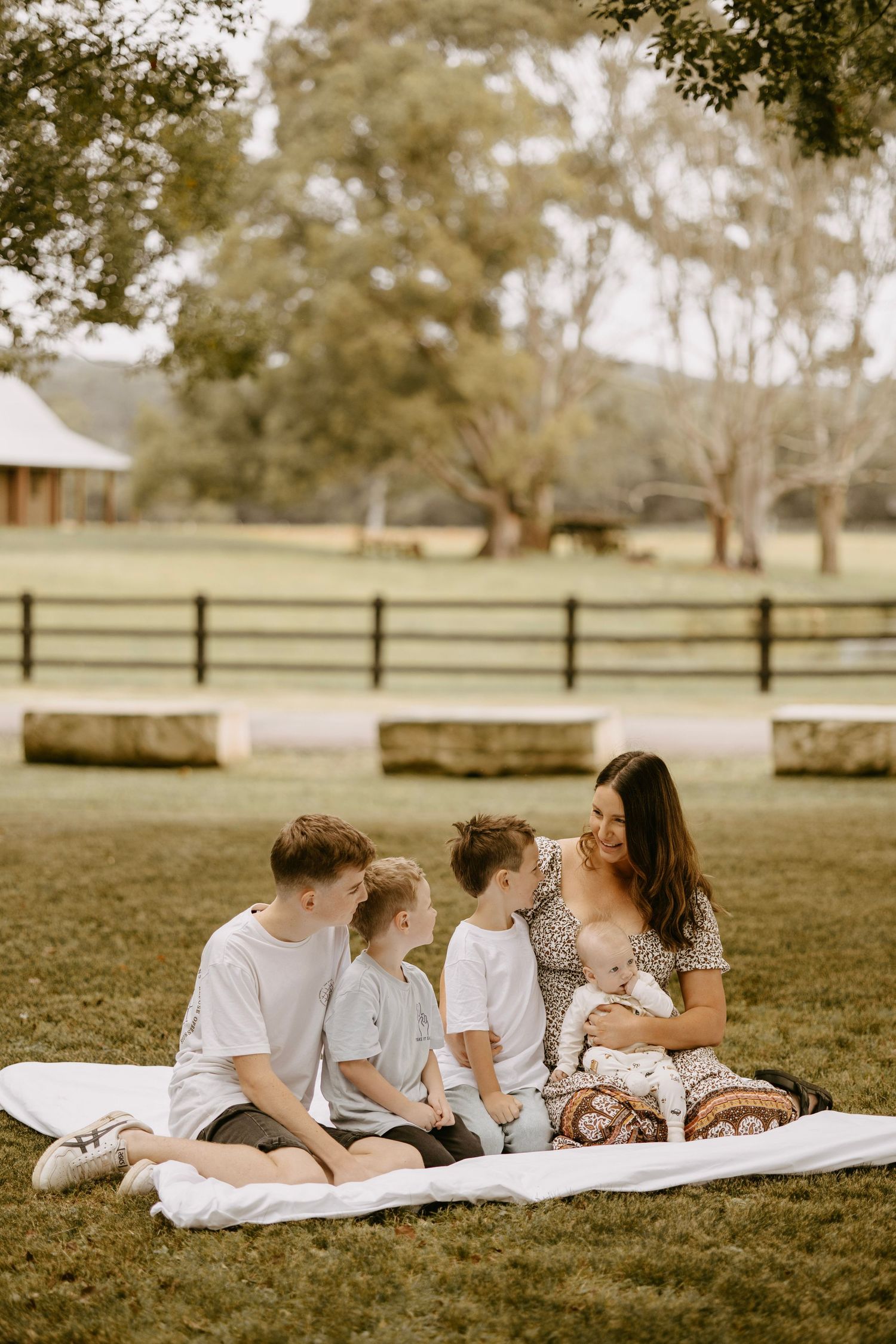 Mother with three children sitting on white blanket in green countryside farm setting.