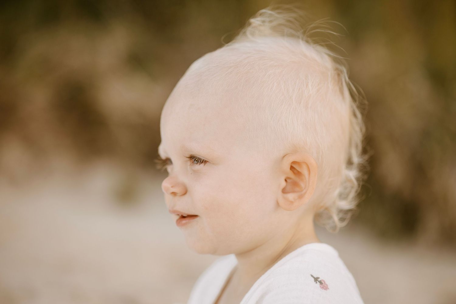 Close-up portrait of blonde baby with wispy hair looking sideways on sunny beach.