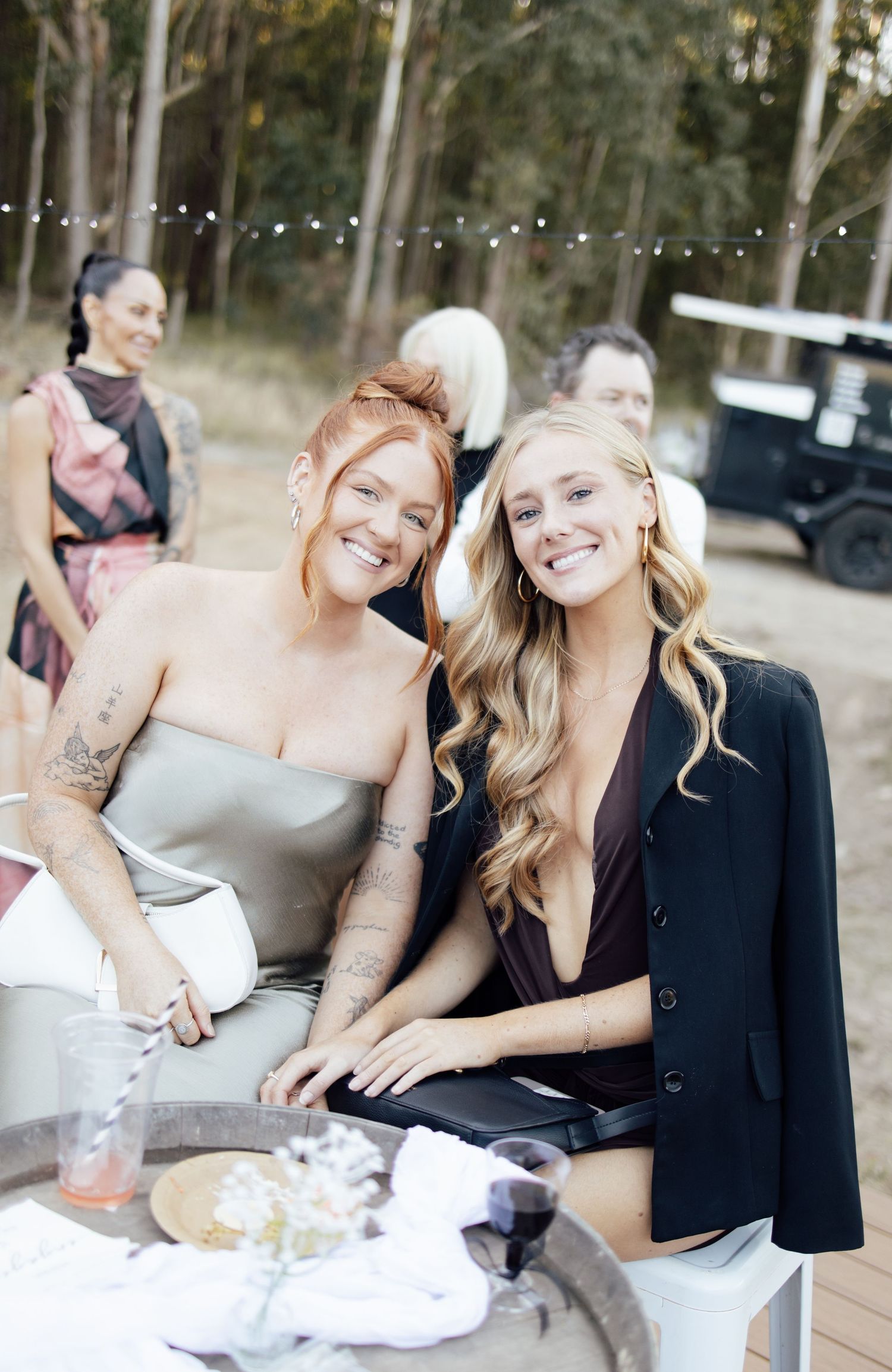 Two smiling women posing together at an outdoor party with string lights and guests in the background.
