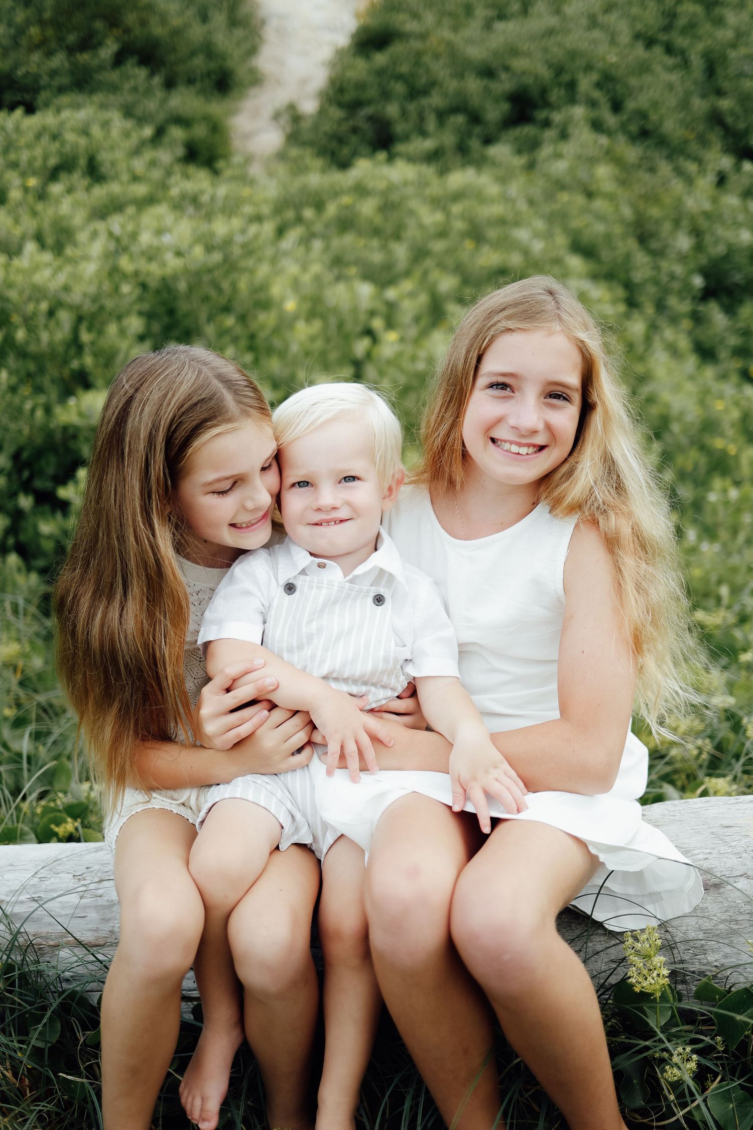 Two sisters in white dresses hugging smiling blonde toddler boy on log outdoors.
