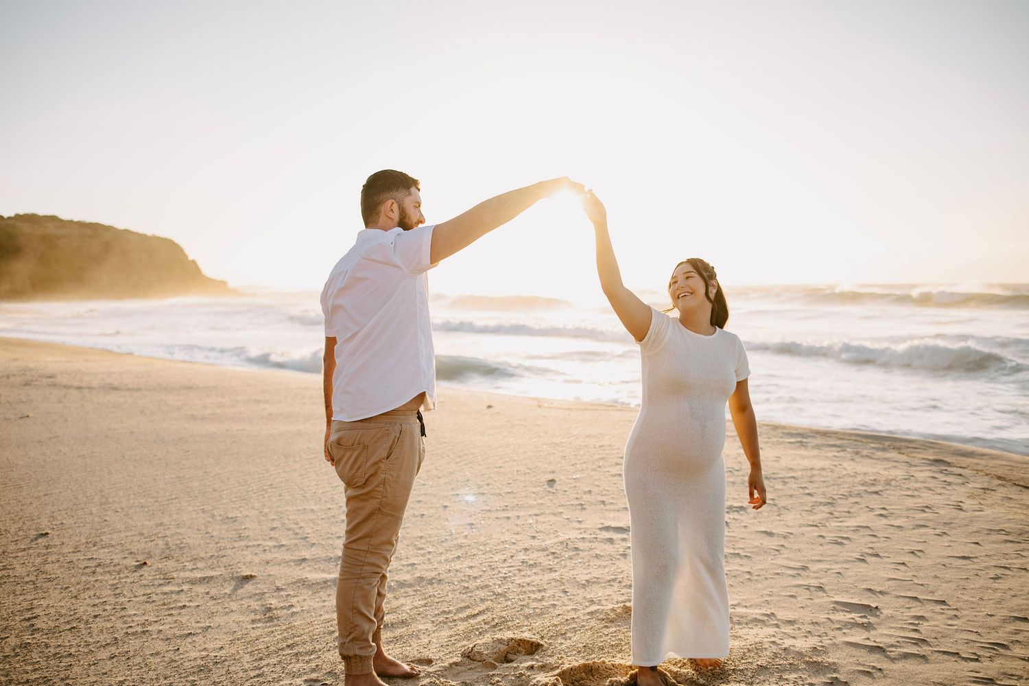 A couple dances on a sunny beach at golden hour, the woman wearing a white maternity dress.