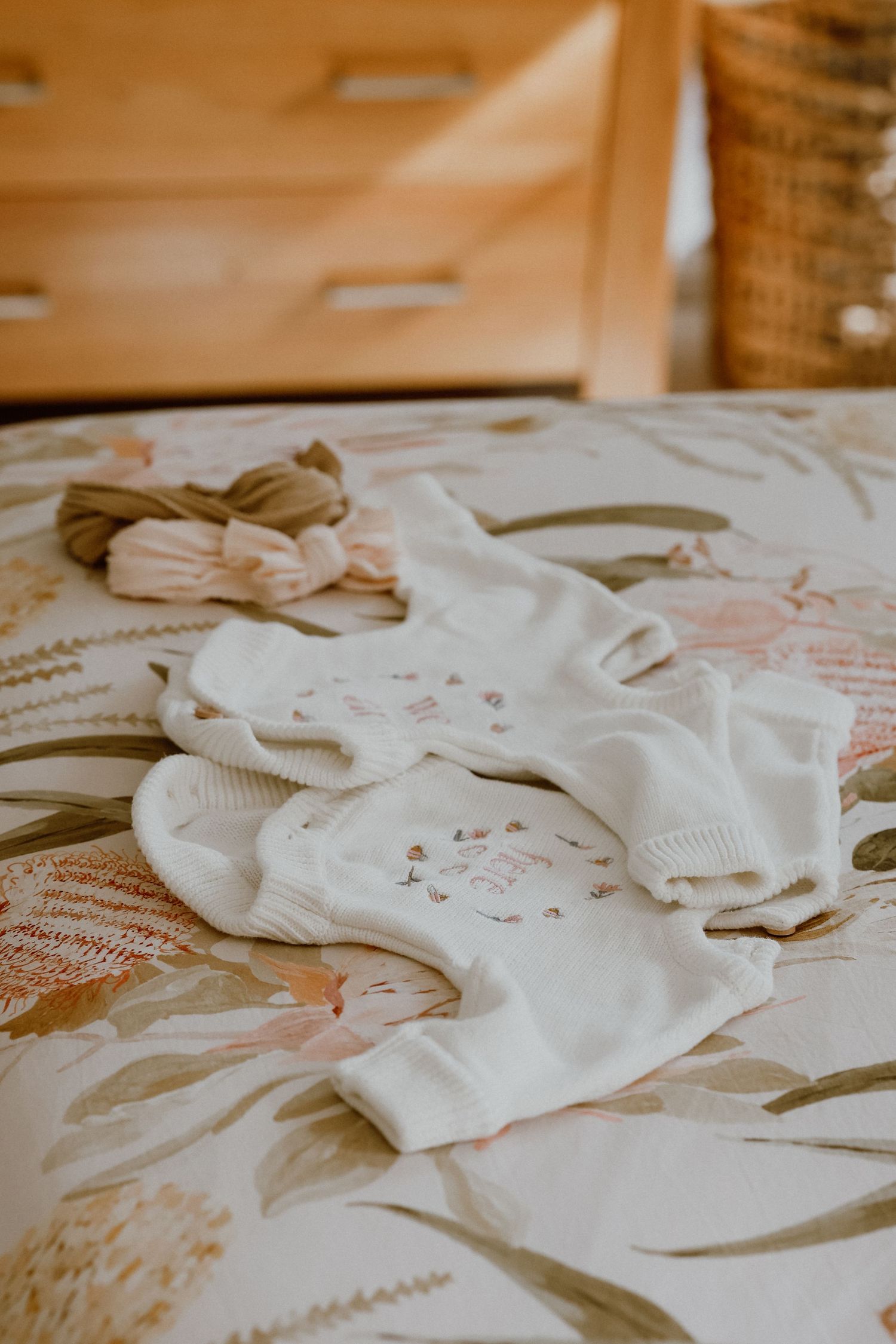 White floral baby onesie and headbands laid out on a floral patterned bed with wooden dresser.
