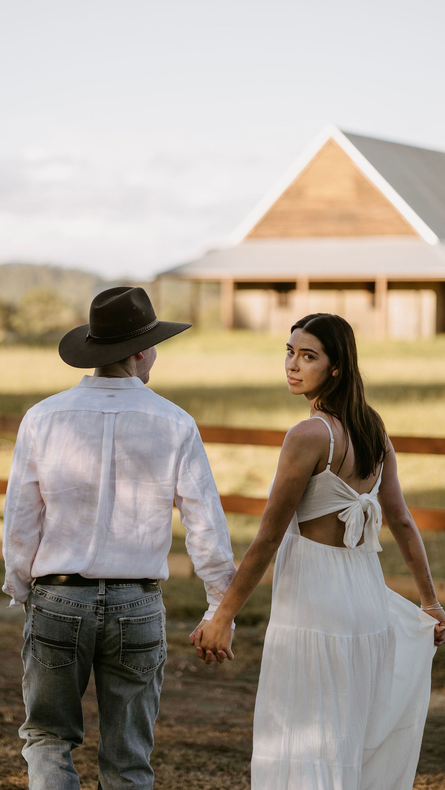 Woman in white tie-back dress looks back while holding hands with man in cowboy hat on farm path.