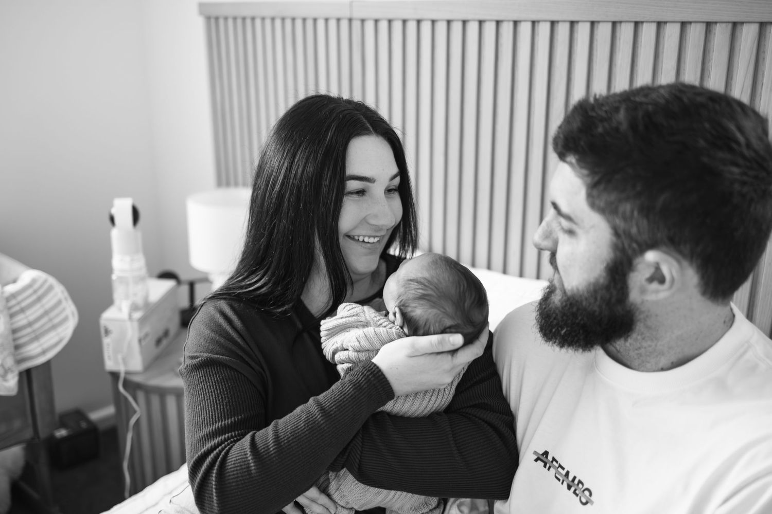 Black and white photo of smiling new parents holding their newborn baby in a hospital room.