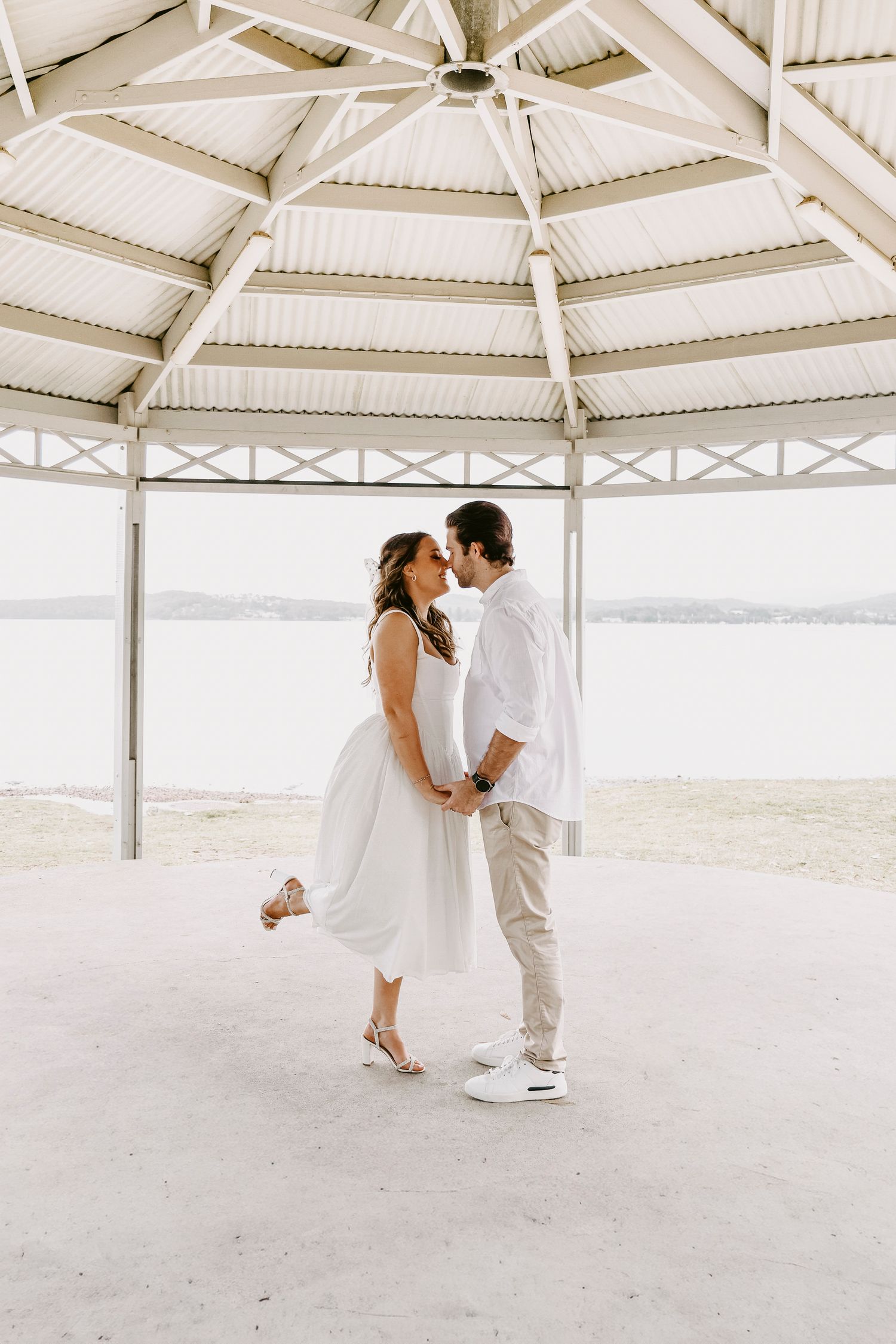 Romantic couple sharing a kiss under a white beachside pavilion with sandy shores behind them.