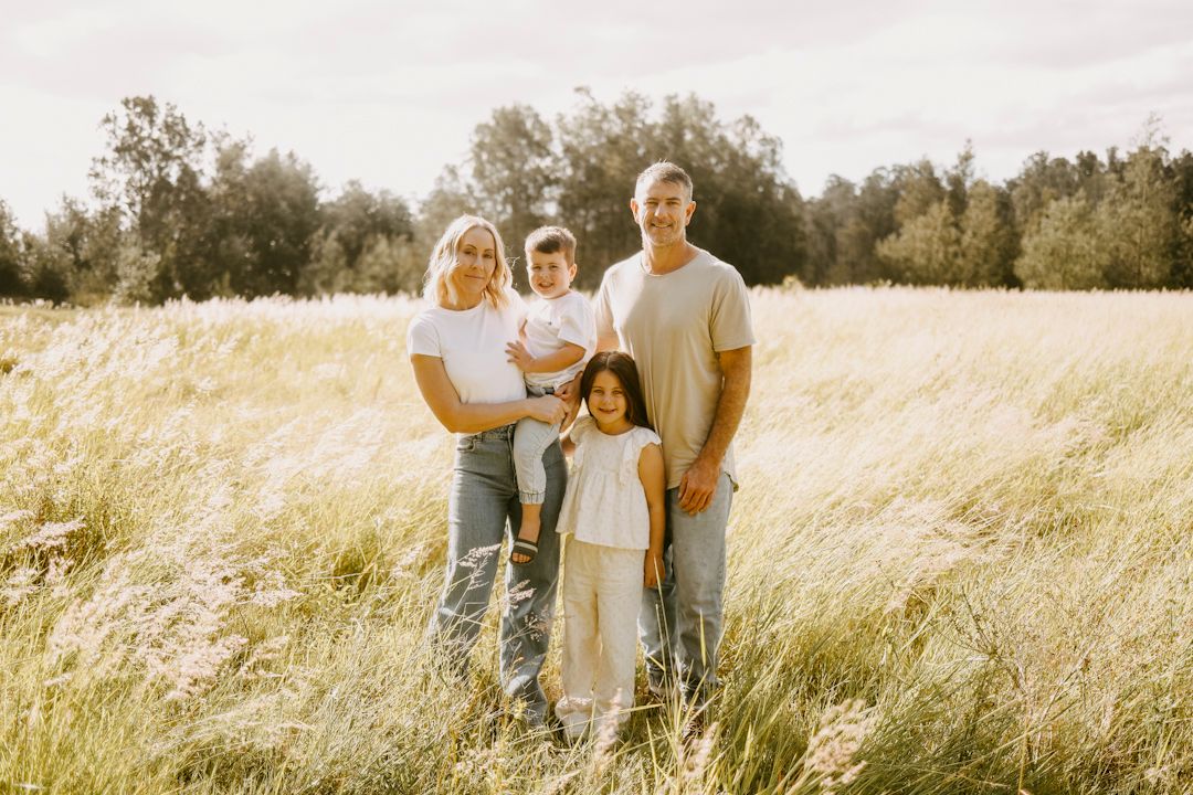 A family of four stands smiling together in a golden sunlit meadow with tall grass and trees.