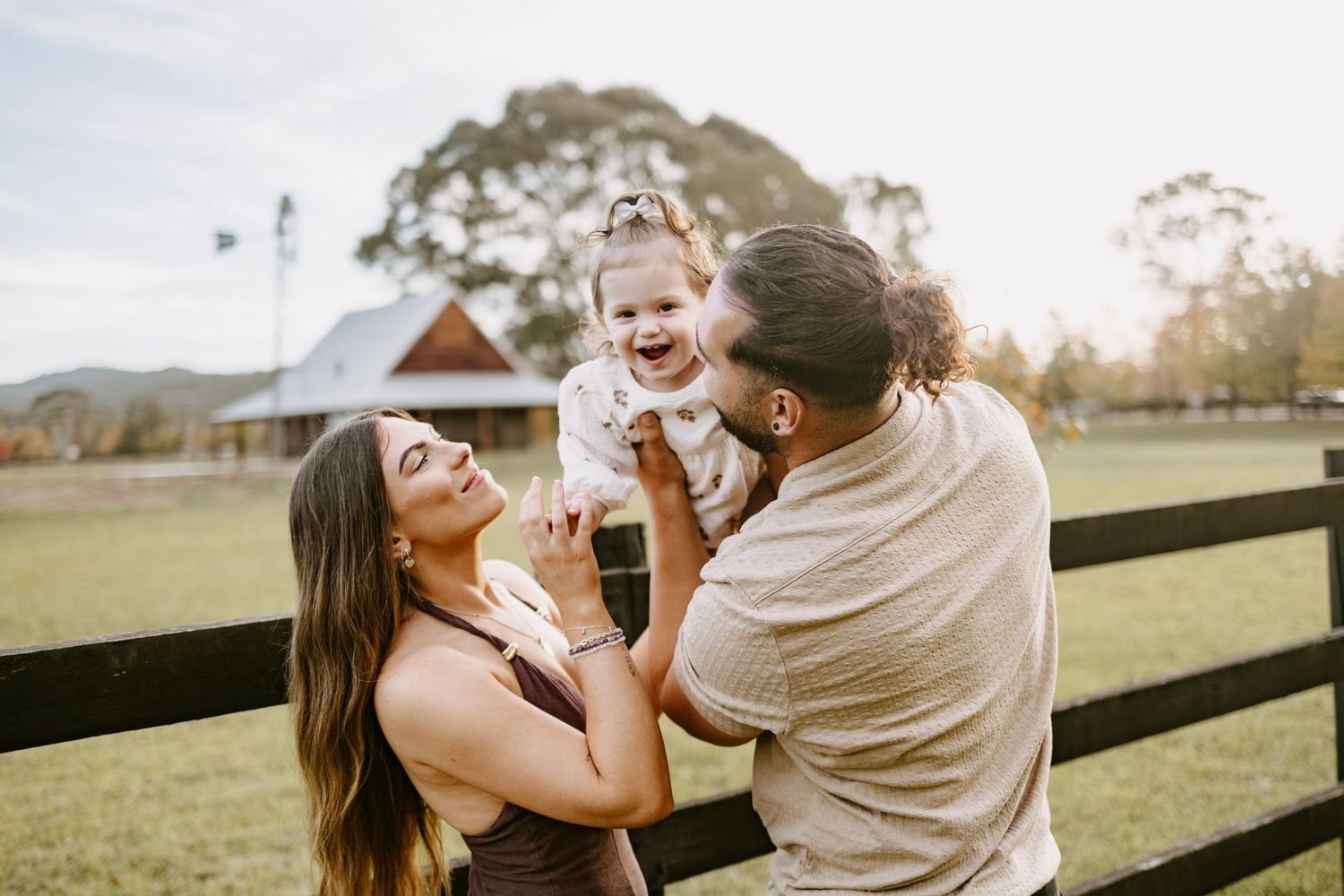 Happy family with parents lifting laughing baby girl outdoors on a farm with green fields.