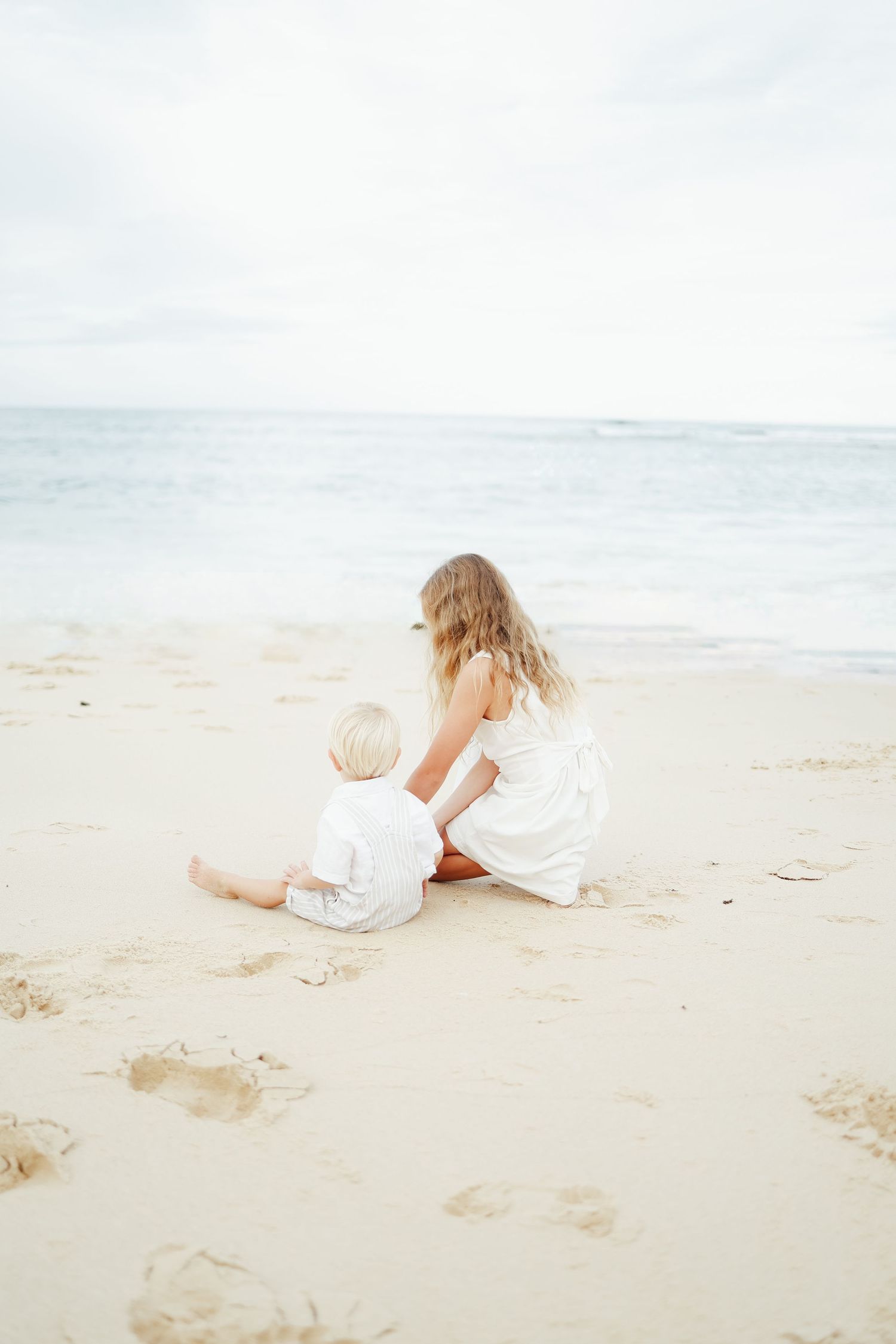 Two young children in white outfits sitting together on sandy beach gazing at the ocean.