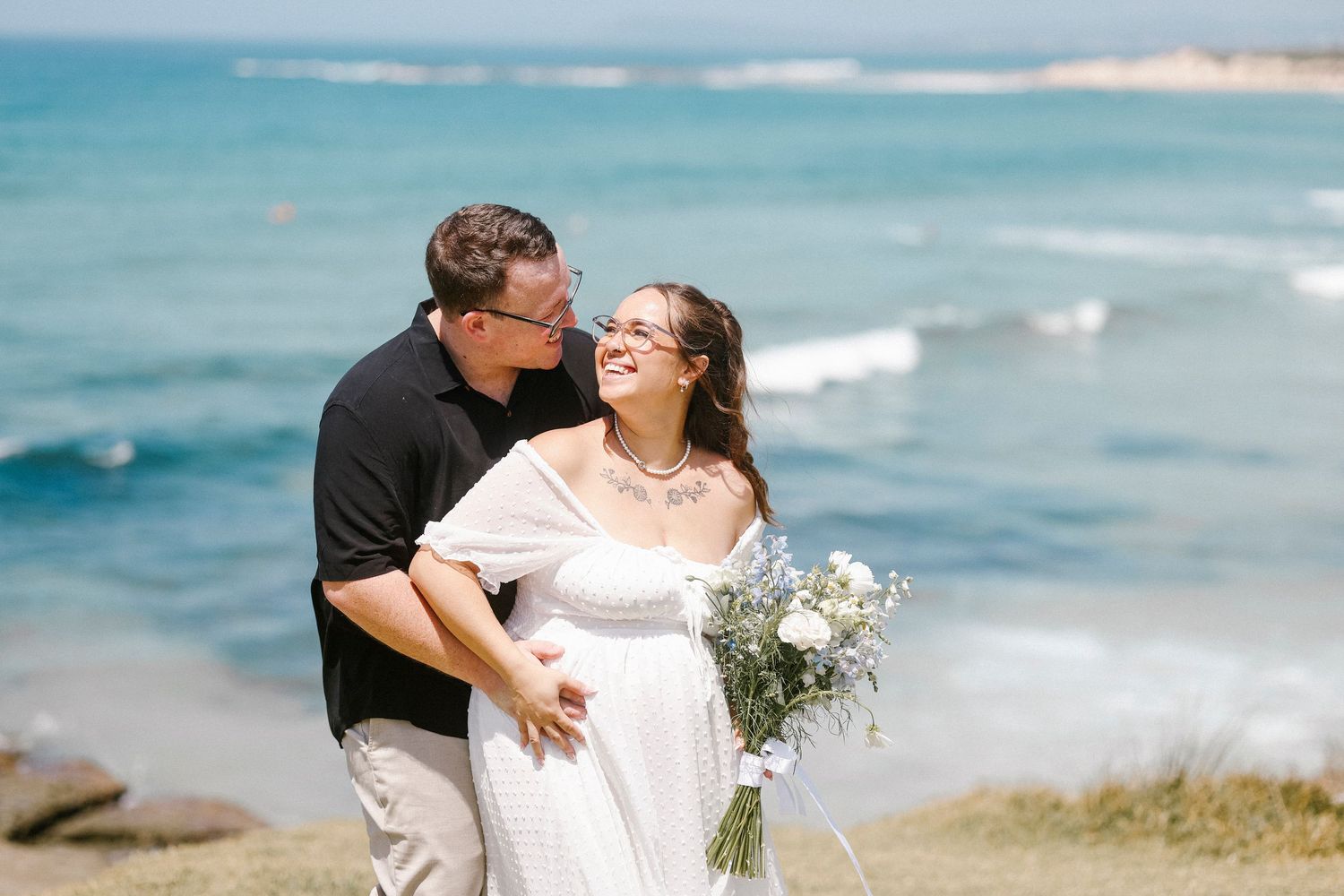 Couple embracing and smiling at each other on a rocky beach with turquoise ocean waves, bride holding white bouquet.