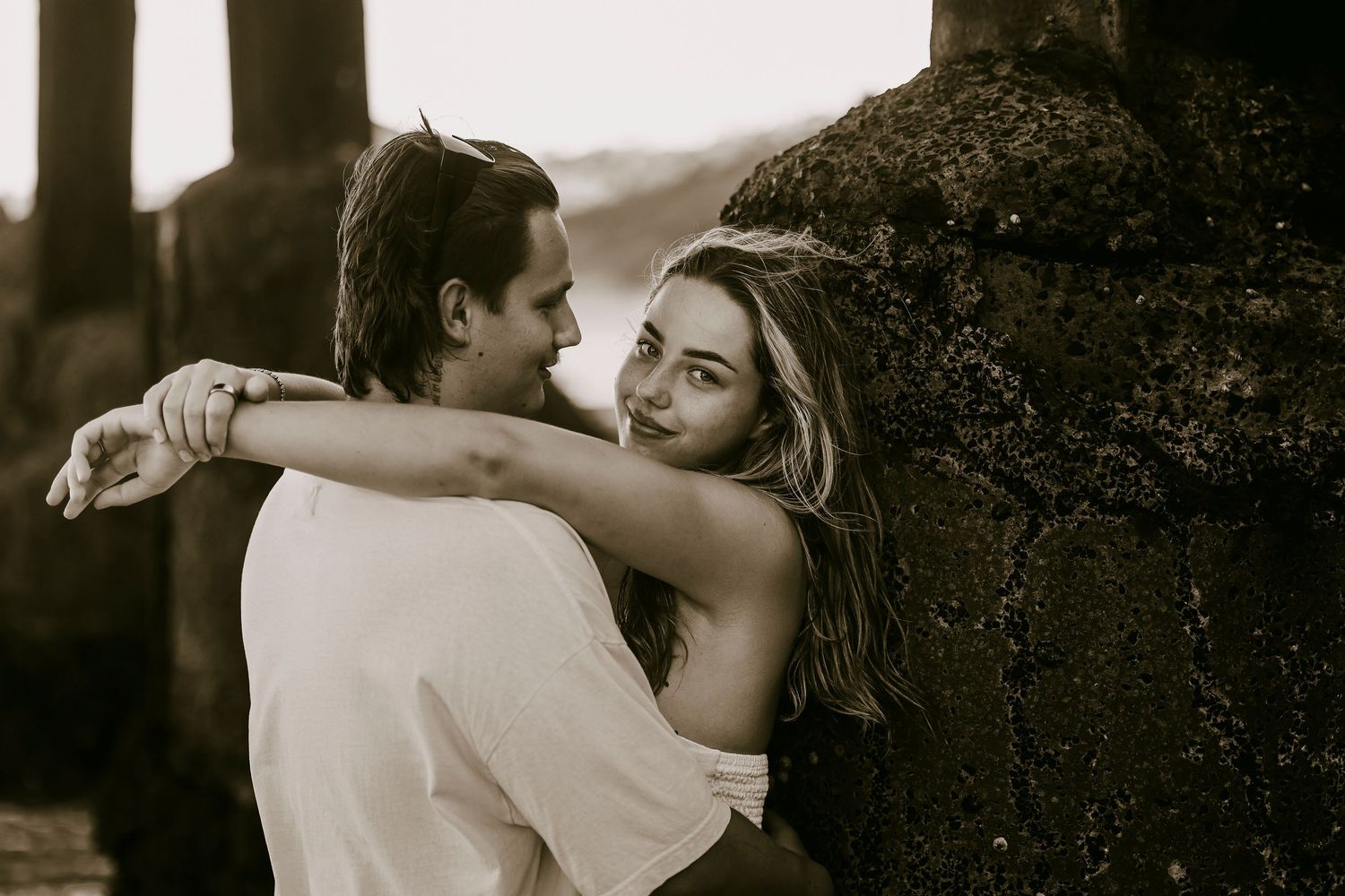Black and white photo of couple embracing near stone wall, woman wrapping arms around mans neck.