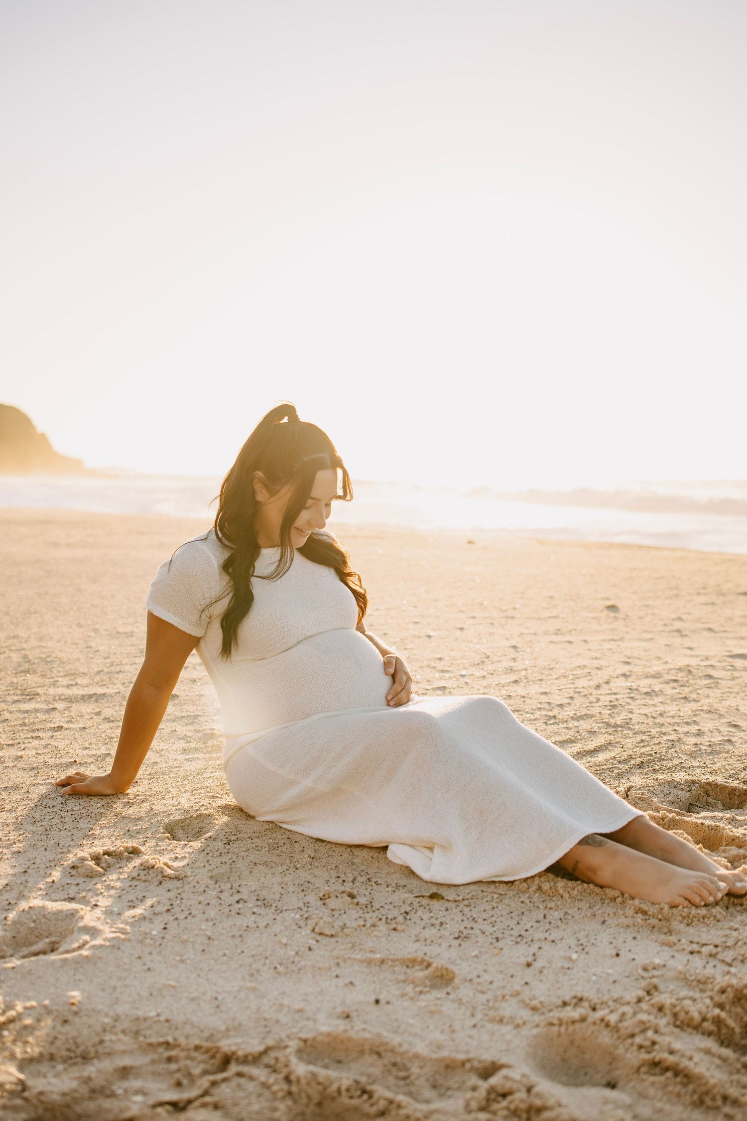 A pregnant woman in a white dress sits barefoot on a sandy beach at golden sunset hour.